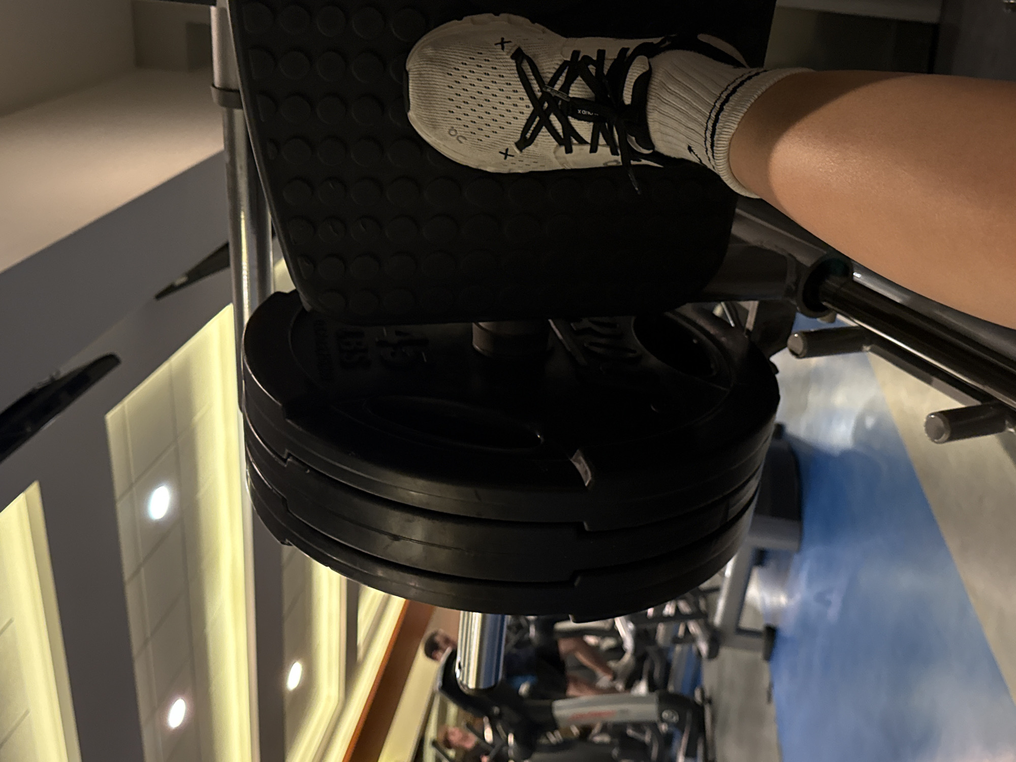 The image presents a close-up view of a person's lower leg and foot performing an exercise on a gym machine, likely a leg press, in a gym located in Mexico City, Mexico. The individual wears a white athletic shoe with black laces, featuring an "On" logo and an "X" mark on its side, along with a white sock with black stripes. Their foot is positioned on a black, textured footplate. The machine is loaded with multiple black weight plates, indicating a resistance exercise. Faint, unreadable text or numbers are visible on one of the weight plates.
The background shows artificial overhead lighting reflected on a dark, polished surface, possibly a ceiling or high wall, creating bright linear patterns. A segment of blue flooring is visible on the right, and indistinct gym equipment is blurred in the farther background. The scene is illuminated by these artificial lights, typical of an indoor fitness facility. No specific time of day or emotions are apparent.
