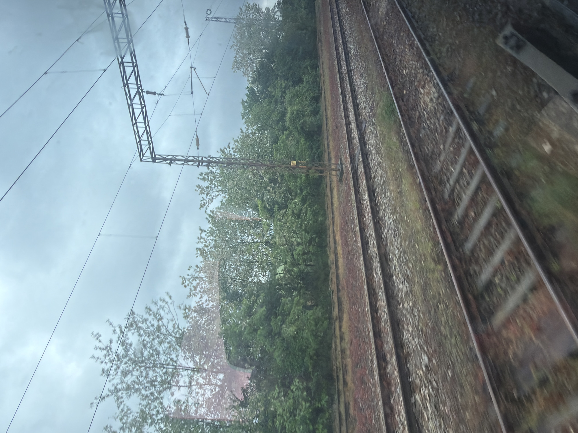 A view from a moving train, showcasing parallel tracks laid with gravel and bordered by lush greenery and trees under a cloudy sky. The composition features an angled perspective capturing railway infrastructure with electrical pylons and cables. The scene is in Pavlikeni, Bulgaria.