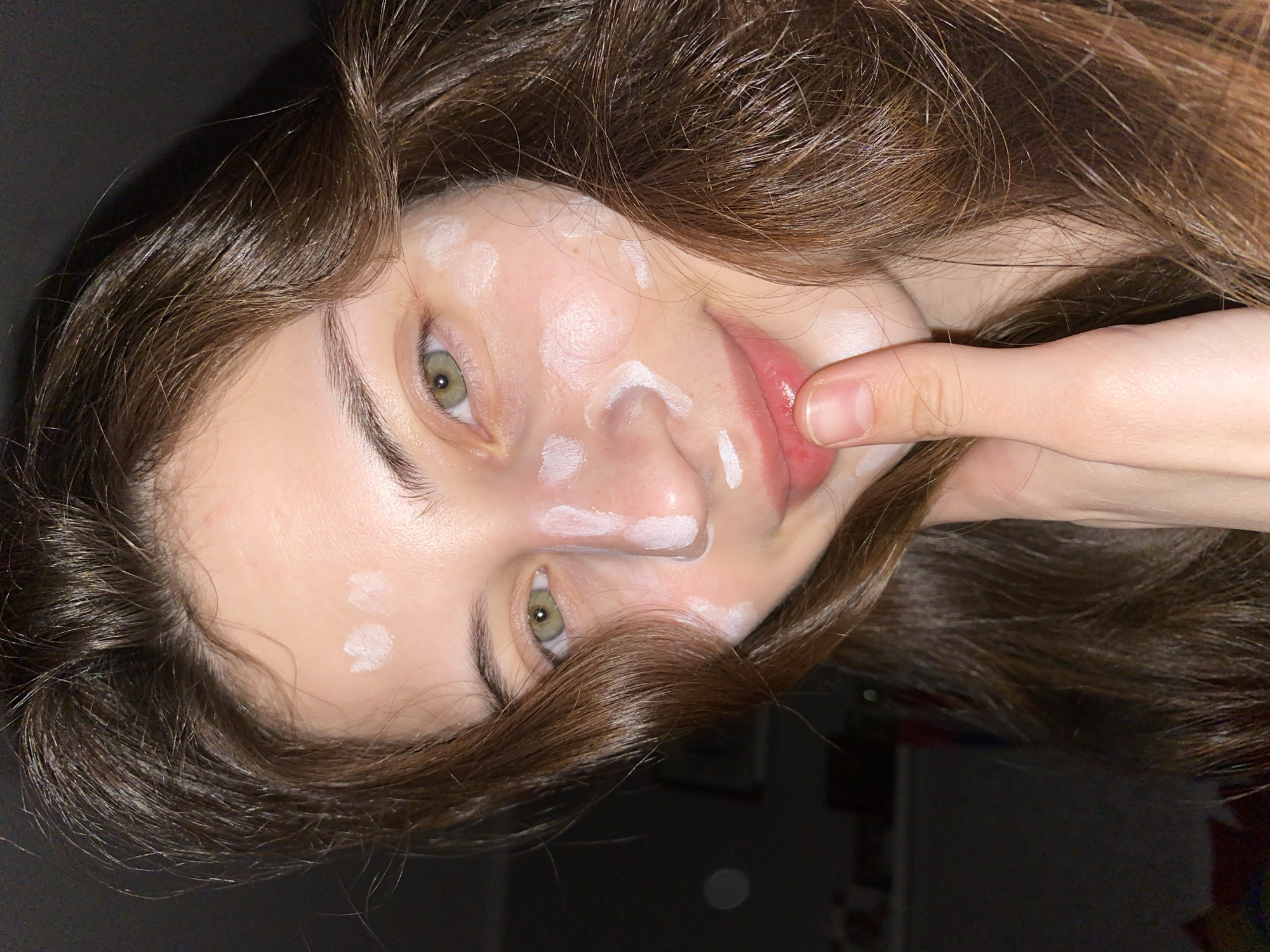 A young woman with light skin, hazel eyes, and brown hair is shown in a close-up shot. She has white cream or lotion applied to her face in small dots or streaks. Her hand is visible, touching her lip. The background is dark and blurry, suggesting an indoor setting. There is no information to indicate that this image was taken in Votkinsk, Russia.