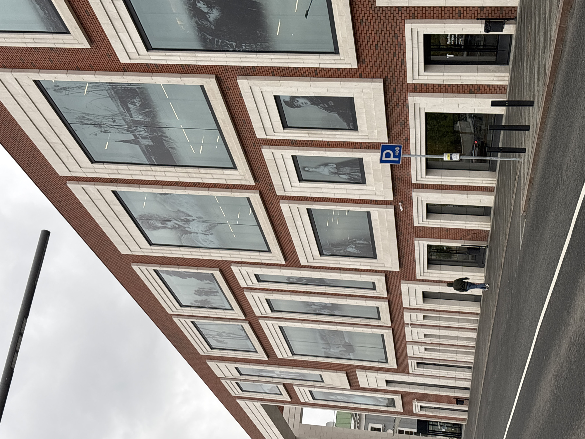The image depicts the facade of a building in Moscow, Russia. The building is constructed with red bricks and features a series of rectangular windows with white frames. Each window appears to have a black and white photograph or image displayed behind the glass.
A parking sign with a blue "P" is visible near the top right of the image. A person is walking along a paved area next to the building. The sky is overcast. A pole or lamppost is visible on the left side of the image. The building appears to be multi-storied with a consistent architectural design repeated across its visible surface.