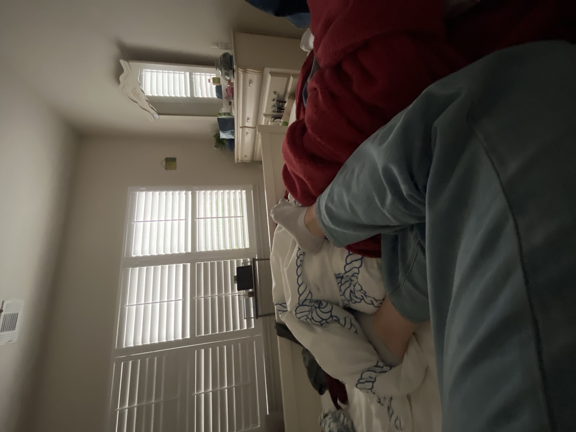 The image shows a cluttered bedroom interior. Two windows with white shutters are visible in the background, allowing natural light into the room. An air conditioning unit is installed on the wall near the left window. A white dresser is positioned against the wall, with various items on top, including what appears to be cleaning supplies. In the foreground, there are piles of bedding, including a red blanket, a white comforter with blue rope patterns, and possibly other items of clothing. A person is in the frame wearing a blue jacket.
There is no information in the image that indicates the location in the city of San Antonio, United States of America.