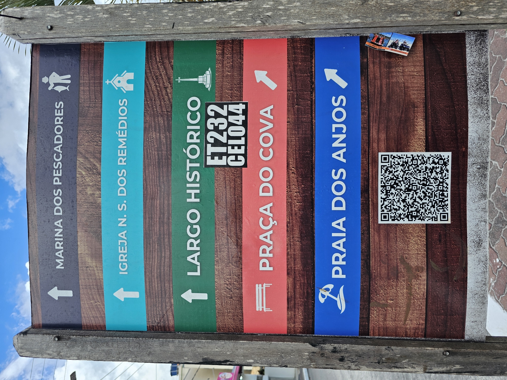 A vertical, rectangular directional sign, framed by two weathered, rough-hewn wooden posts, stands outdoors in Arraial Do Cabo, Brazil. The sign features five colored panels, each indicating a local landmark. From top to bottom:
1. A dark gray panel reads "MARINA DOS PESCADORES" with an upward arrow and an icon of a person holding a fish.
2. A teal panel reads "IGREJA N. S. DOS REMÉDIOS" with an upward arrow and a church icon.
3. A green panel reads "LARGO HISTÓRICO" with an upward arrow and a monument/cross icon. A black sticker with white text "ET232 CEL044" is affixed over the center of this panel.
4. A red panel reads "PRAÇA DO COVA" with a right-pointing arrow and a bench icon.
5. A blue panel reads "PRAIA DOS ANJOS" with a right-pointing arrow and an icon of a beach umbrella/palm tree.
Below the directional panels, a large black and white QR code is displayed on the brown, wood-grain patterned background of the sign. A small, folded brochure or sticker, displaying images of what appear to be boats or diving activities, is partially affixed to the right wooden post, slightly overlapping the lower right section of the sign's main face.
The sign is situated on a paved surface composed of reddish-brown interlocking pavers. In the background, under a partly cloudy blue sky, indistinct elements of an urban environment are visible, including power lines, a utility pole, and a building with a light-colored facade.