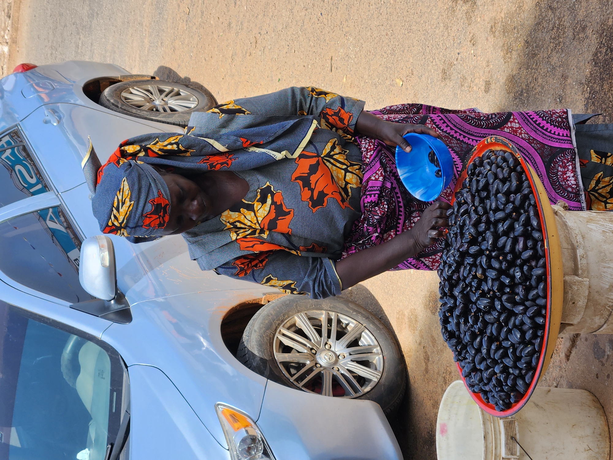 An overhead, outdoor shot captures a person engaged in a transaction next to a silver car. The individual, dressed in a vibrant, patterned outfit featuring a headwrap and top with large orange and yellow leaf designs on a dark blue fabric, and a skirt with a pink and black geometric pattern, is positioned in the center-right of the frame. Their hands are busy, scooping dark, oval-shaped fruit, resembling plums or large olives, from a large, reddish-rimmed bowl into a smaller blue measuring bowl.
The large fruit bowl rests on two stacked white buckets, which serve as a makeshift stand. A silver sedan is parked closely on the left side of the frame, revealing its front headlight, side mirror, and a wheel with a visible Toyota logo on its hubcap. The ground beneath is a light brown, suggesting dirt or asphalt. The scene is brightly illuminated by natural daylight, indicating it is daytime. Reflected text, appearing as "12A" in reverse, is faintly visible on the car's window. The image portrays an everyday street vending scene.