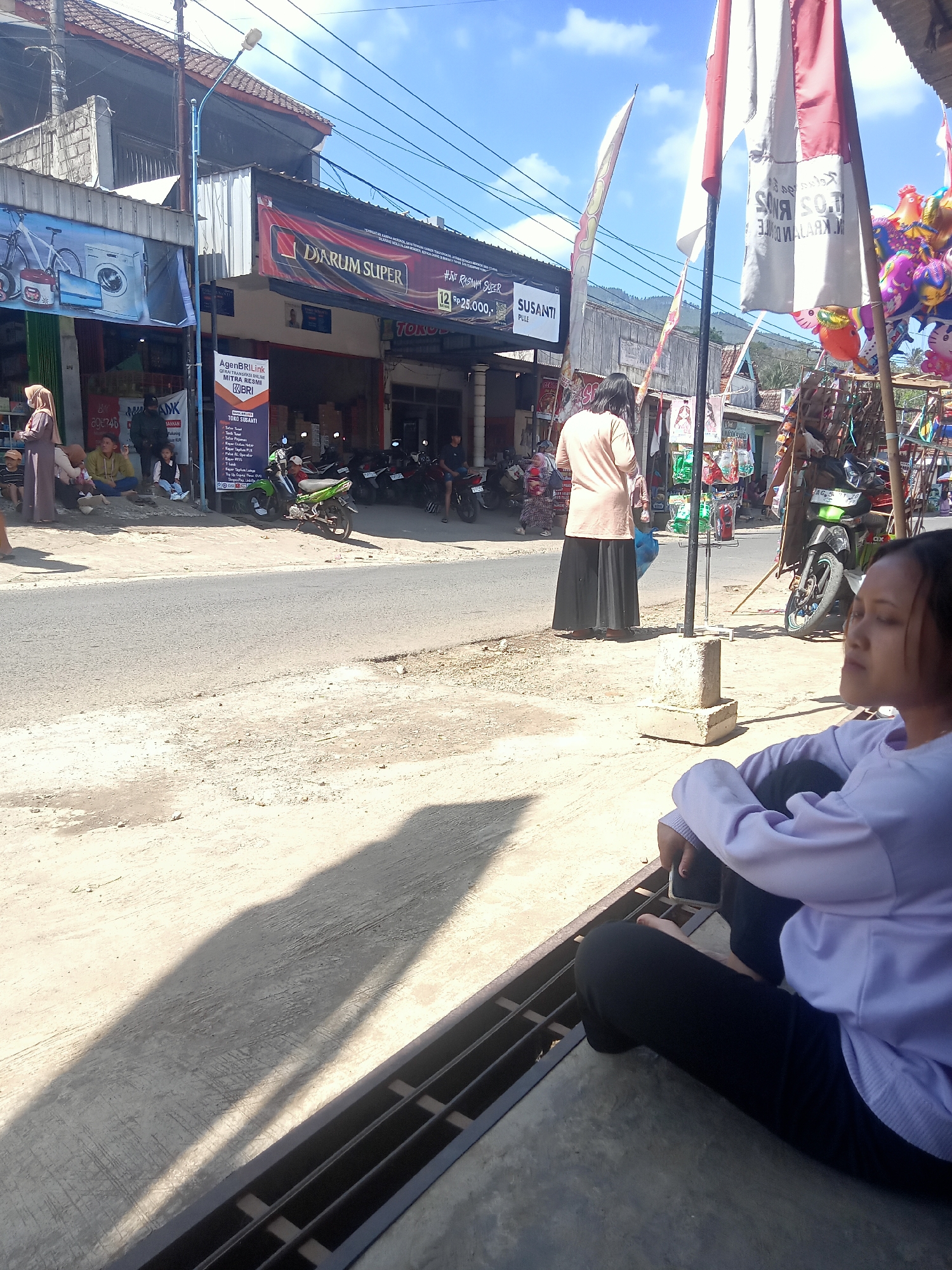 The image depicts a street scene in Pule, Indonesia, likely on a sunny day, with a clear blue sky and scattered clouds. In the foreground, a young woman is sitting on the ground, facing away from the camera and to the right, with her legs crossed and her arms resting on her knees. She is wearing a light purple sweatshirt and black pants. A shadow of a person is cast on the ground to her left.
The middle ground shows a street with several parked motorcycles. A woman in a tan shirt and long black skirt is standing on the sidewalk, facing the street. Several other people are seated on the sidewalk further down the street, some appearing to be in conversation. Shops and businesses line the street, with signs indicating various establishments, including "DARUM SUPER," "AGENBRIlink MITRA RESMI BRI," and "TOKO SUSANTI." Flags, including what appear to be red and white Indonesian flags, are visible. Colorful balloons are displayed on the right side of the image.
The background features buildings, power lines, and a hillside with some vegetation. The overall environment suggests a typical commercial area in a town or village in Indonesia.