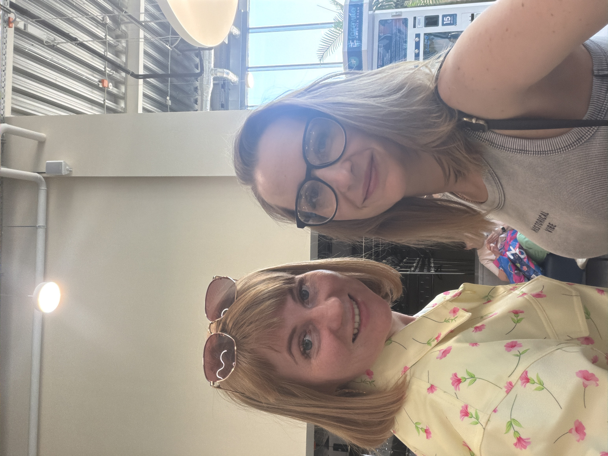 Two women are taking a selfie indoors. The woman in the foreground has short, light brown hair, sunglasses pushed up on her head, and is wearing a yellow floral-patterned shirt. The woman in the background has shoulder-length blonde hair and is wearing black-rimmed glasses and a grey tank top. The background includes a glimpse of a vending machine, a bright interior, and what appears to be a clothing rack with colorful garments. There is no indication that the location is in the city of Moscow, Russia.