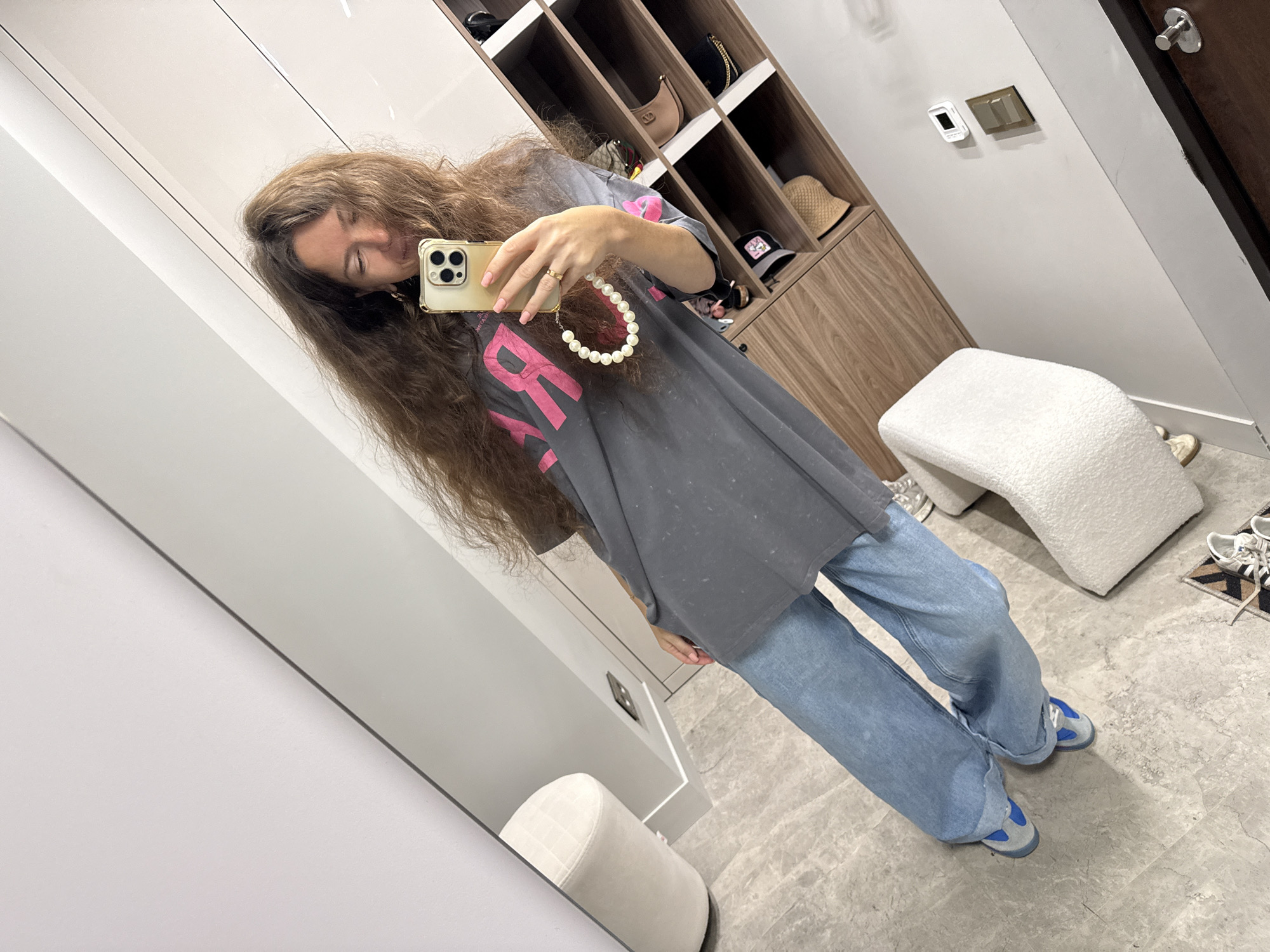A woman is taking a full-body mirror selfie in what appears to be a modern dressing room. She is wearing a dark gray oversized t-shirt with a bright pink graphic and a pearl necklace. Her lower half is clad in light blue, wide-leg jeans, and she's wearing blue and gray sneakers. Her long, brown hair is flowing around her.
In the background, there is a large mirror reflecting the scene, a wooden shelving unit filled with various items like hats and bags, and a plush white armchair. The flooring is a light-colored tile. The overall aesthetic suggests a well-organized, contemporary space. The specific location in Dolgoprudny, Russia, cannot be determined from the visual information provided.