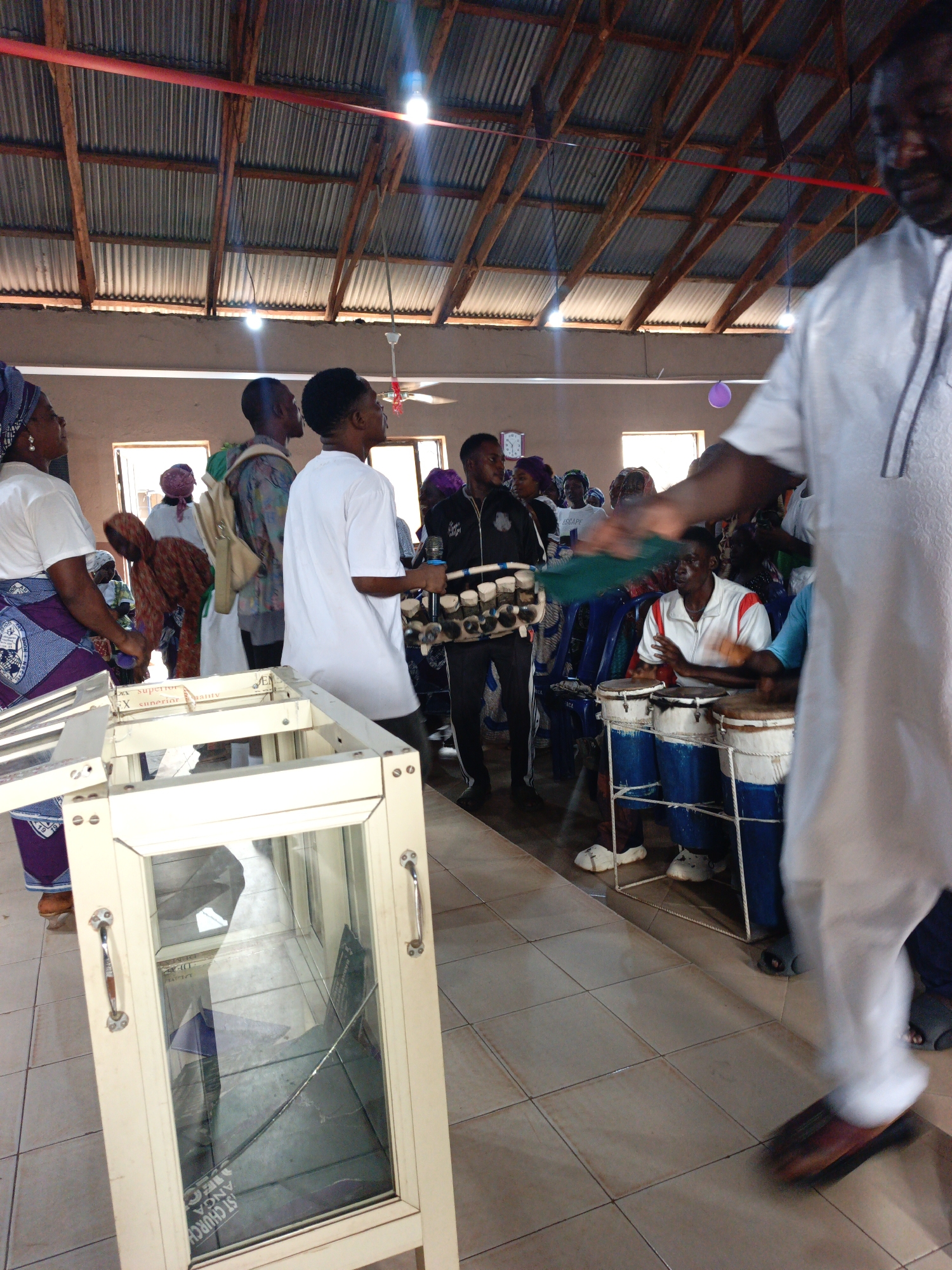 The image captures an indoor scene in what appears to be a church in Akwanga, Nigeria. The building has a corrugated metal roof supported by wooden beams. Several individuals are present, with varying degrees of focus. On the left, some people are wearing traditional African attire. A person in a white shirt carries a microphone rack. In the center, a musician is playing a set of drums. An open glass-fronted structure stands on the left. A man wearing a white robe is moving in the foreground. The floor is tiled. Natural light enters through windows along the walls. A single, purple balloon hangs in the upper background.