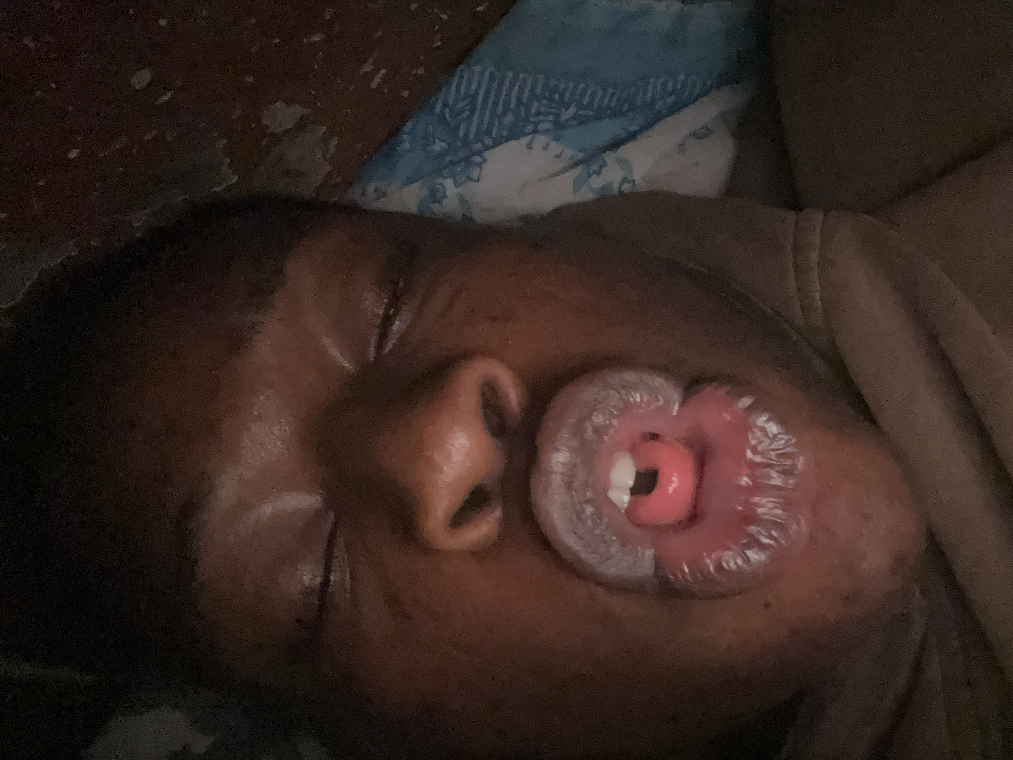 A close-up, high-angle photograph shows a person sleeping. The person has dark skin, a closed eye, and full lips. The person's mouth is slightly open, revealing a single white tooth and part of their tongue. The person appears to be lying on a dark surface with a blue-and-white patterned pillow behind them. The location is Unknown, Unknown.