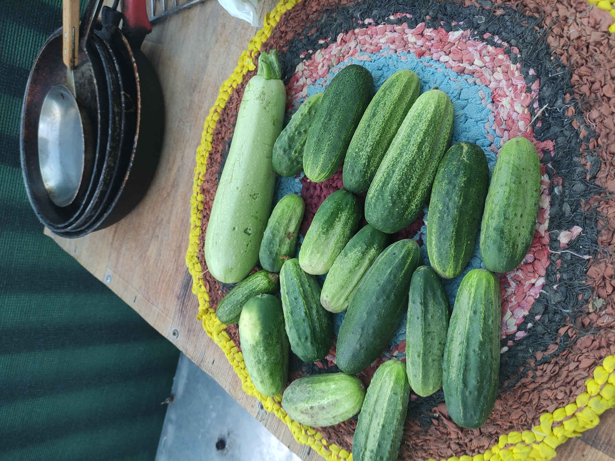 A close-up, high-angle outdoor shot shows a collection of fresh produce, specifically cucumbers and one large zucchini, arranged on a colorful, circular woven mat. The mat, featuring shades of yellow, brown, red, and blue, rests on a wooden surface, likely a table. In the background, a dark green corrugated surface is visible, with a stack of three dark metal frying pans resting on the wooden surface to the right. The overall scene suggests a harvest or preparation for cooking. The image was captured in Rybnitsa, Moldova.