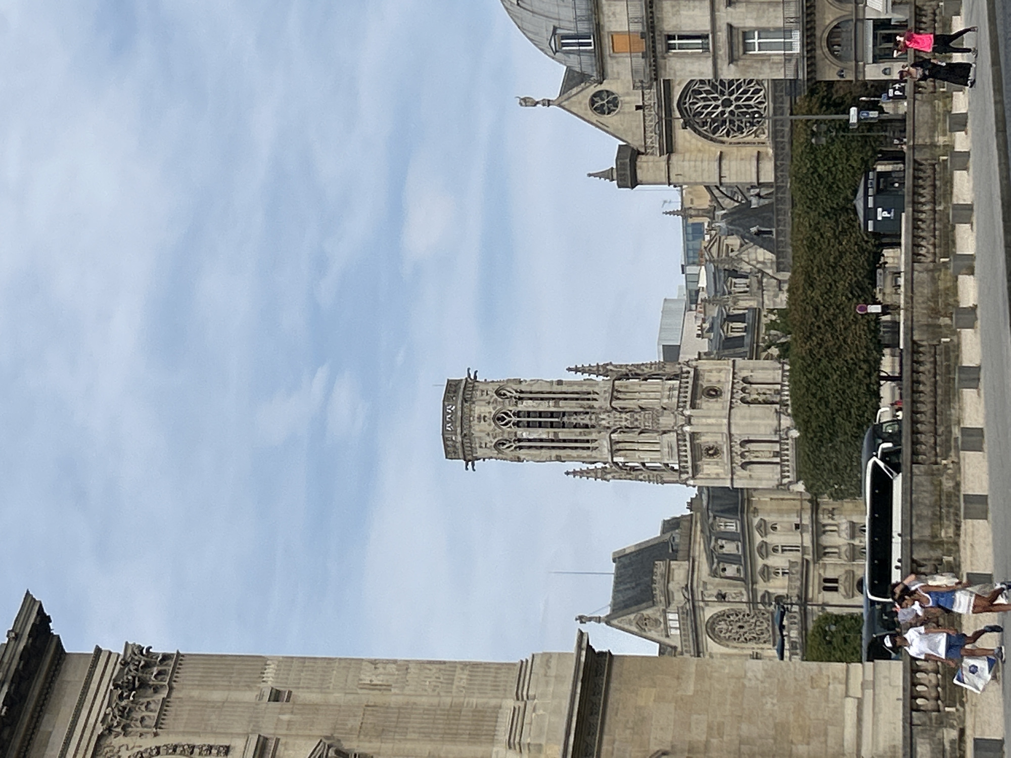 This is a vertical, eye-level, full shot image of the Sainte-Chapelle in Paris, France, on a slightly overcast day. The perspective is from a street level, looking across a body of water towards the iconic landmark.
In the foreground, on the left, a large stone building with classical architectural details and fluted columns dominates the left side of the frame. The stonework appears weathered. A sidewalk and a stone barrier with square concrete bollards separate the street from the building.
Further back, a tall, ornate Gothic tower with intricate tracery and spires rises prominently from the center of the frame. This is the bell tower of Sainte-Chapelle. To the right of the tower, the roof of the Sainte-Chapelle with its domed section and stained glass windows can be seen. Adjacent buildings with similar stone architecture and arched windows are also visible, forming a cityscape.
In the mid-ground, a white bus with tinted windows is parked or moving slowly. A dense green hedge runs along the street behind the barrier. A small, dark green kiosk-like structure with a visible "P" logo is situated in front of the hedge. Streetlights and traffic signs are also present.
On the sidewalk in the mid-ground, several individuals are walking. Two young people, a male wearing a white polo shirt and blue shorts, and a female in a blue and white dress, are walking from left to right, carrying a large flat object. Further to the right, two women are walking from right to left, one in a long black dress and the other in bright pink sportswear.
The sky is a pale blue with scattered, wispy white clouds. The overall lighting suggests a daytime scene with diffused sunlight.