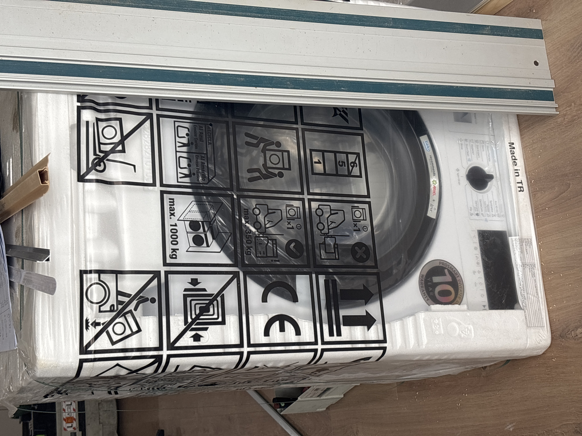 The image shows a new, packaged Beko washing machine with various warning symbols printed on its plastic wrapping. The machine is white with a black rubber seal around the drum opening. A control panel is visible with the "Made in TR" (Turkey) mark. The packaging includes a "10 Year Warranty" sticker and symbols indicating how the machine should be handled (e.g., "do not use a trolley"). The machine is positioned on a wooden floor, possibly in a home or storage setting. A piece of aluminum or white metal trim is visible in the top part of the photo. There's no information in the image to indicate that the location is Plovdiv, Bulgaria.