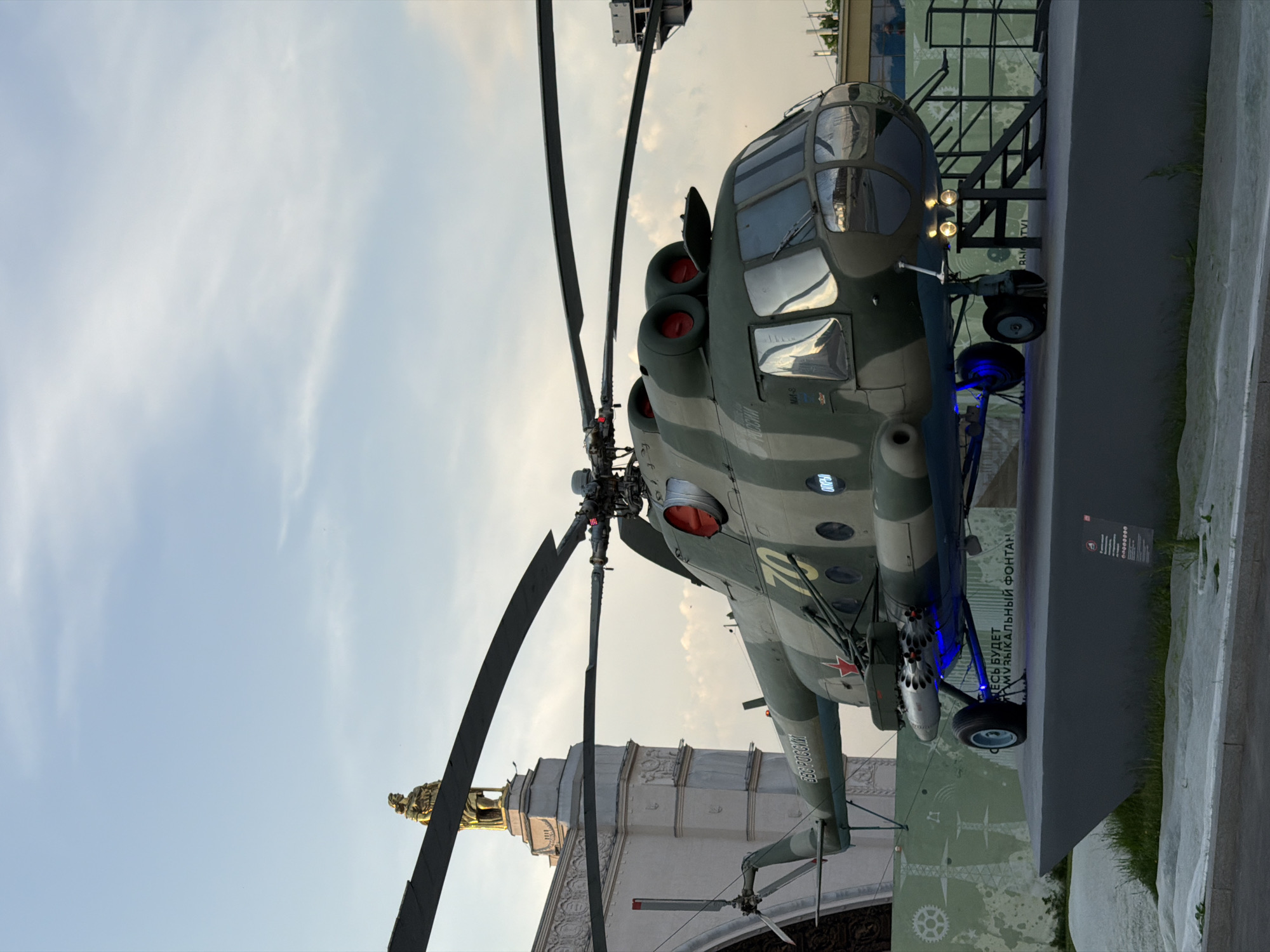 The image shows a Russian Mil Mi-24 Hind attack helicopter on display. The helicopter is painted in a camouflage pattern and has a red star on its fuselage. The helicopter is on a raised platform. In the background is the Friendship of Nations fountain, a famous landmark of the All-Russia Exhibition Centre (VDNKh) in Moscow, Russia.