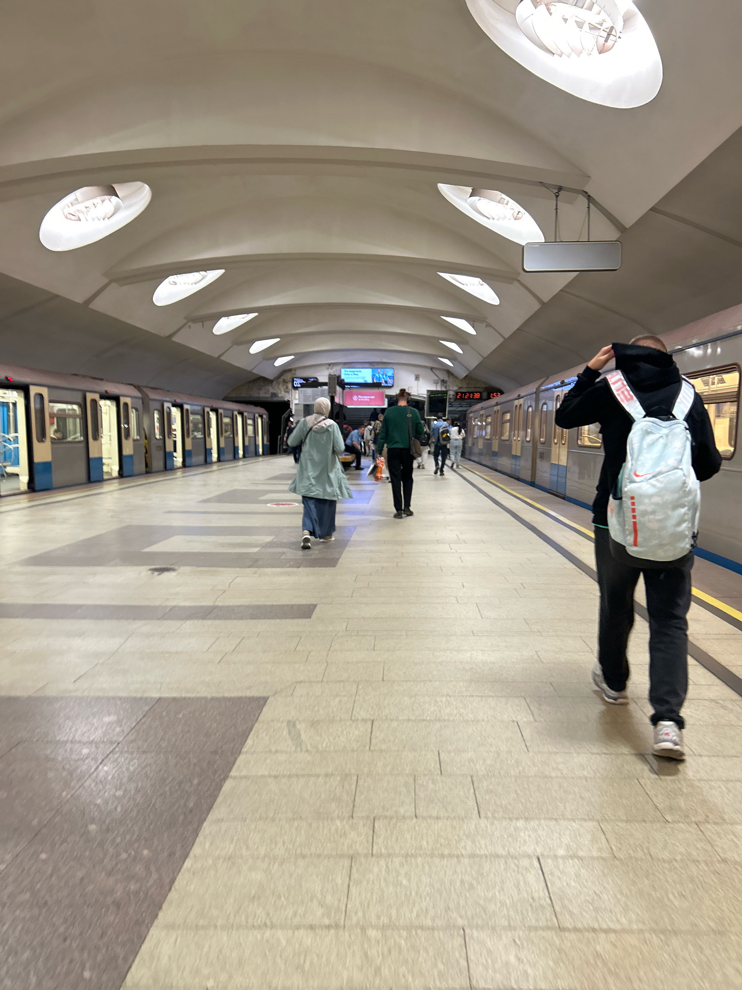 The image depicts a well-lit subway station platform in Moscow, Russia. The architecture features a curved, white ceiling with recessed oval lights. The platform itself is paved with light-colored rectangular tiles, with a darker tiled area in the foreground. A train with open doors is visible on the left side of the platform.
Several individuals are present. In the foreground, a person wearing a black hoodie and dark pants is walking away from the camera, carrying a light blue and white patterned backpack. To the left and further down the platform, a person wearing a light blue jacket and dark pants, with a headscarf, is also walking away. In the middle ground, a person in a green long-sleeved shirt and dark pants is walking towards the train. Other individuals are scattered further down the platform, some appearing to be waiting for the train.
The overall atmosphere suggests an active transit hub during the day. The lighting is bright and even, illuminating the spacious station. The perspective is from the platform looking down its length.