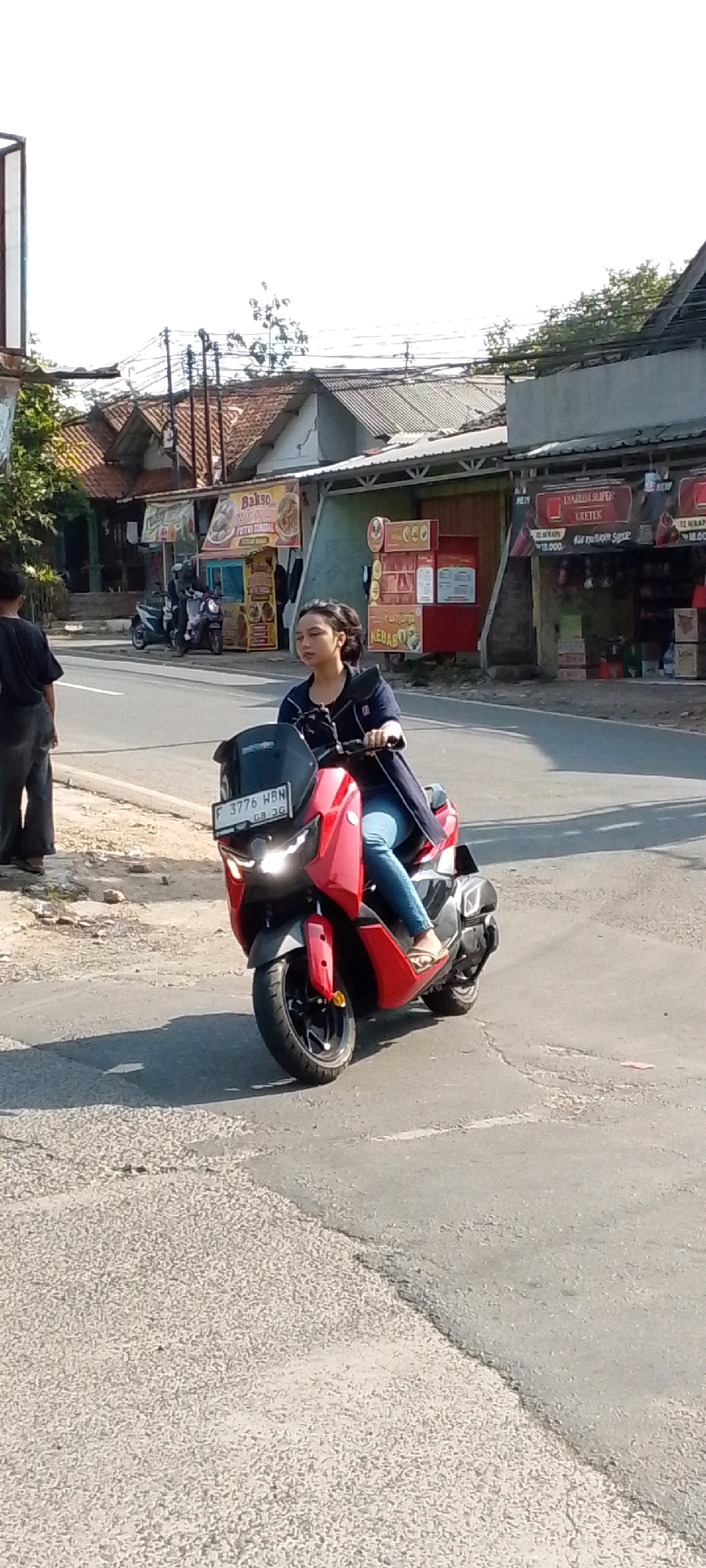 A young woman with dark hair is riding a red and black scooter on a paved road. She is wearing a dark blue jacket and blue jeans, with her feet in flip-flops. Her left hand is on the handlebar, and her right hand is not visible. The scooter has a black windshield and a license plate that reads "F 3776 HBN GB 3G". She is looking forward, slightly to her right. The road is partially shaded, with a rough, gray asphalt surface. To the left of the road is a dirt embankment with some scattered gravel. In the background, there are several buildings with tiled roofs, some of which appear to be shops with signs advertising food items like "Bakso" and "YumYum Kebab". Overhead, there are power lines and trees. The sky is bright and clear, indicating daytime. The scene appears to be an outdoor street environment in a town or city, likely in Indonesia given the license plate format and architectural style.