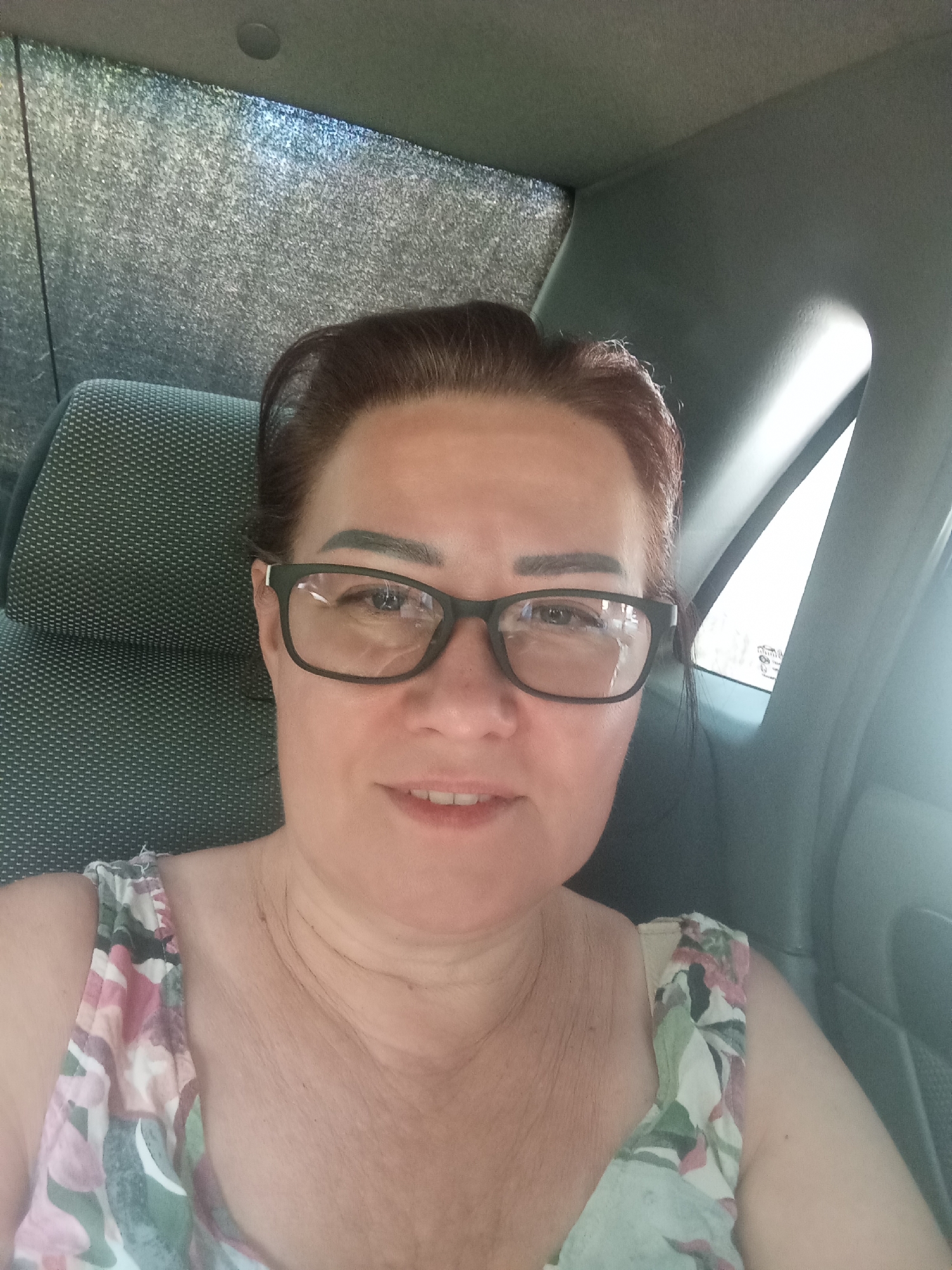 A medium shot shows a woman with brown hair and glasses, wearing a floral-patterned top, sitting in the back seat of a car. The car's interior is visible, with a textured seat and a gray headrest. The background includes a reflective surface on the left and a window with light streaming through on the right, suggesting an outdoor environment. The scene appears to be in Yangikurgan, Uzbekistan.