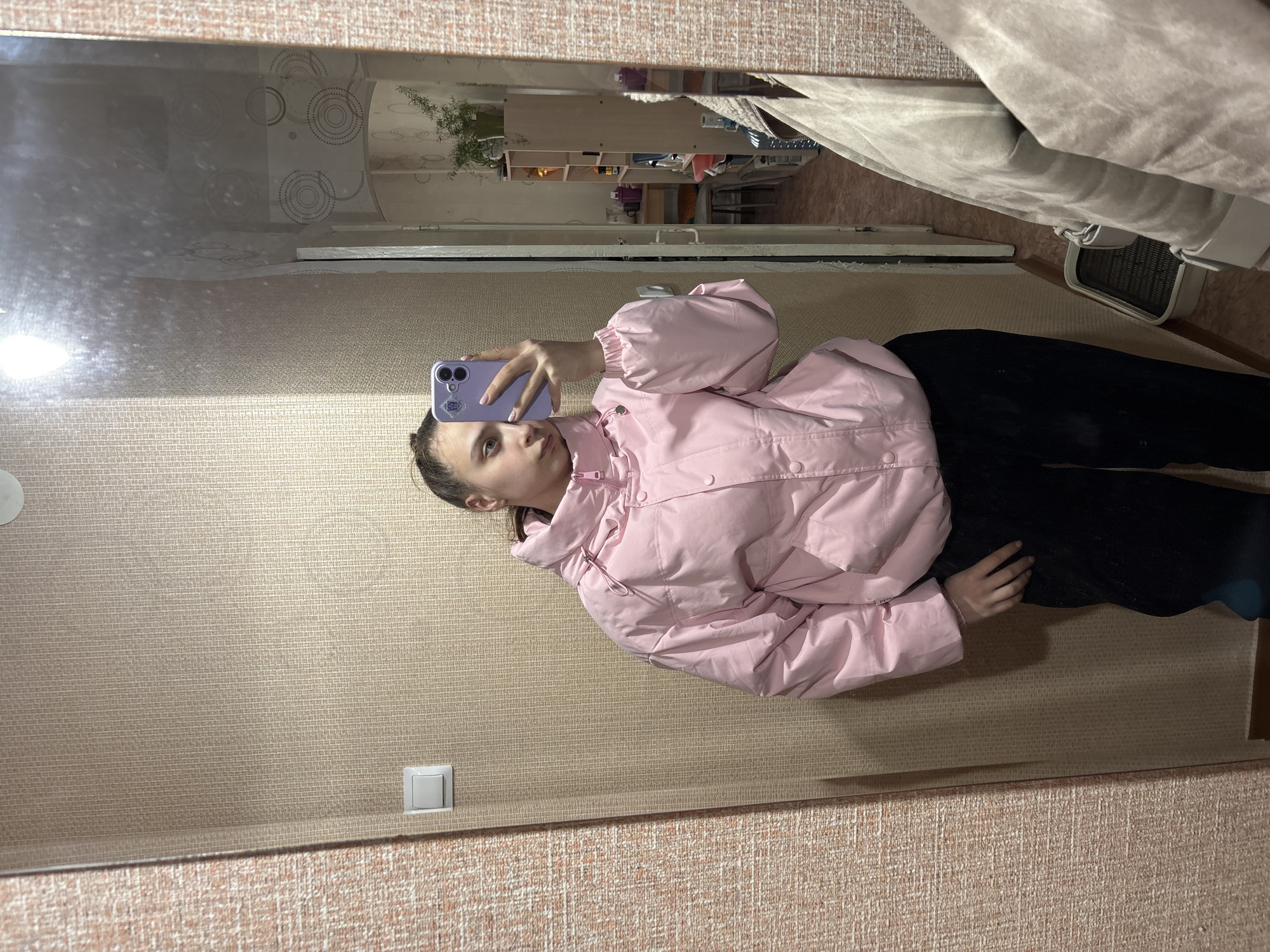 A young person, appearing to be female, is captured in a mirror selfie within an indoor setting in Chaikovsky, Russia. She is wearing a light pink puffer jacket and black pants or a skirt. Her hair is pulled back, and she holds a light purple smartphone with a matching case in front of her, angled to capture her reflection. A small, indistinct sticker is visible on the phone case, and faint text, possibly "ICM" or "HCM," is visible on the sleeve of her jacket. Her expression is neutral, as she looks towards the phone screen.
The room reflected in the mirror features textured light brown or beige wallpaper and a white light switch on the wall. In the background of the reflection, elements of a domestic space are visible: shelves containing various items, a potted houseplant, and what appears to be a bed or sofa covered with light grey, textured fabric. A floor fan is also partially visible on the floor near the furniture. The lighting suggests an indoor environment, likely under artificial illumination.