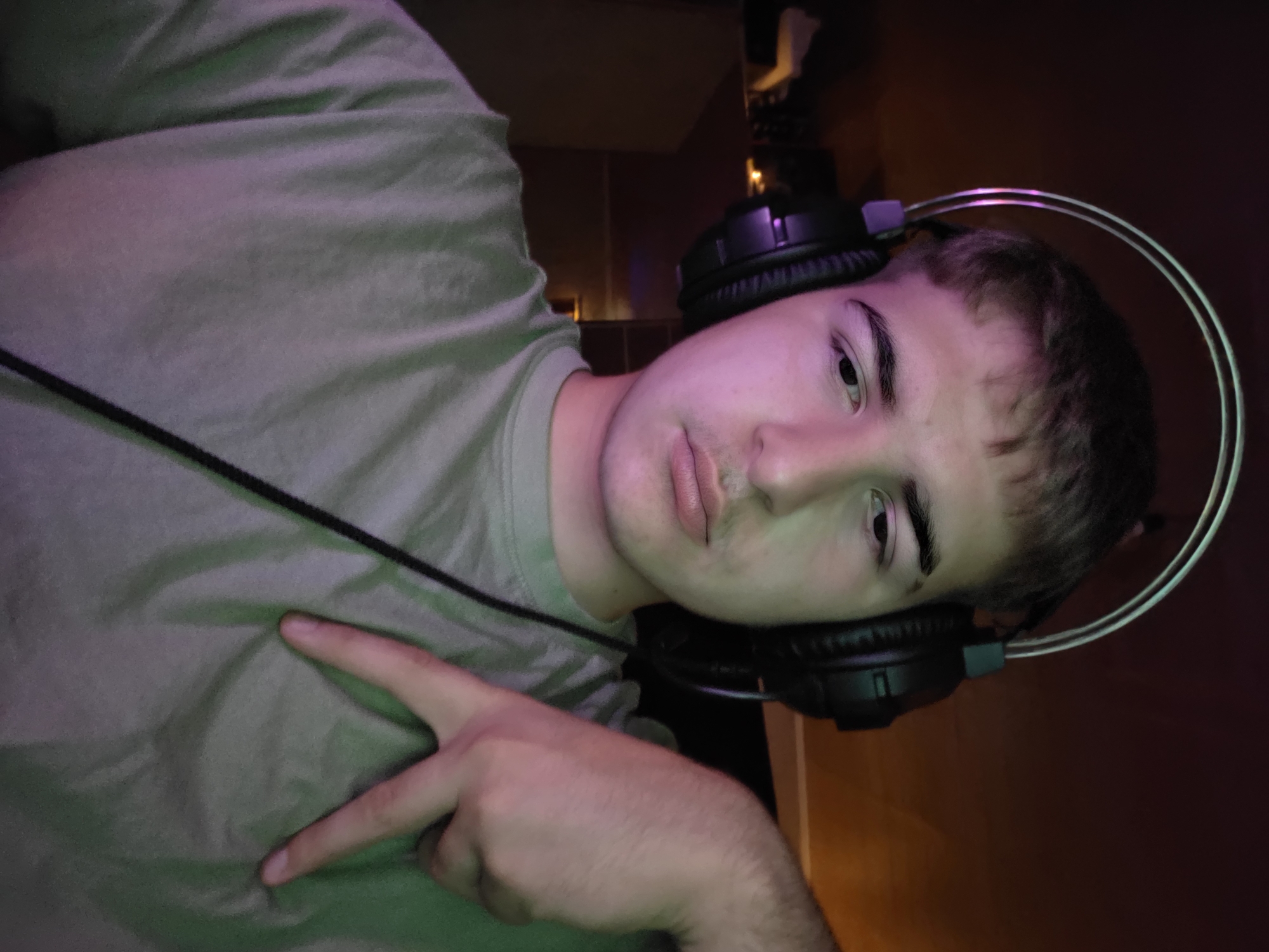 A young male wearing headphones and a light green t-shirt is depicted in a selfie. He is looking directly at the camera with a neutral expression. His left hand is raised, with his index and middle fingers extended in a peace sign, and his hand is resting on his t-shirt. The background is dark, with some faint lighting and what appears to be shelving or furniture. The image is illuminated with purple and green lighting, suggesting an artificial lighting setup. This image was taken indoors, likely in a personal space such as a room. There is no discernible information in the image to indicate its location in the city of Aleksandrovo, Bulgaria.