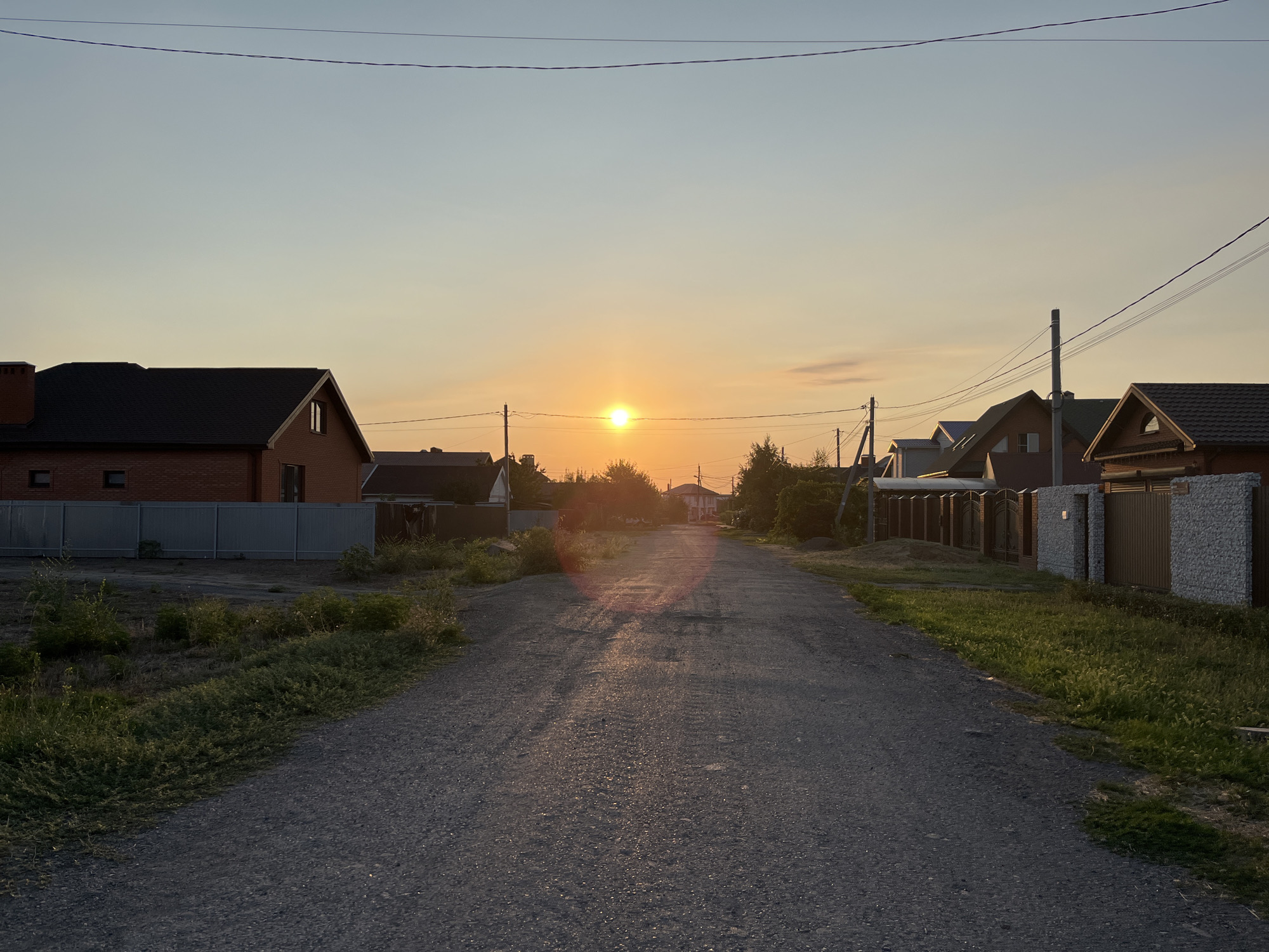 This is a medium-shot, eye-level photograph taken in the city of Taganrog City District, Russia, during what appears to be sunset. The scene depicts a gravel road stretching into the distance, lined with houses and fences on either side. The sun, a bright orange orb, is visible low on the horizon, casting a warm glow and creating lens flare on the road. The sky is a pale blue fading to a pale orange near the horizon. Overhead, electrical wires are strung across the frame. The houses appear to be residential, with various architectural styles and fencing. The vegetation on the sides of the road consists of grass and some shrubs, which are bathed in the soft light of the setting sun. There are no people visible in the frame. The overall impression is one of a quiet, rural or suburban street at dusk.