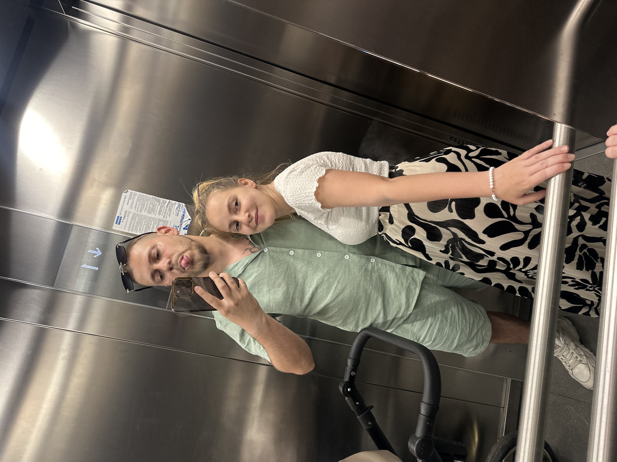 In a metallic elevator, a man and a woman pose for a mirror selfie. The man, wearing a light green short-sleeved shirt and matching shorts, sticks his tongue out while holding a smartphone. He has sunglasses perched on his head. The woman, in a white textured top and a black and white floral skirt, smiles at the camera. A stroller is visible to the left of the man. The elevator walls are reflective, showing the interior and the occupants. The floor of the elevator is dark grey. A few informational posters are affixed to the elevator wall.
