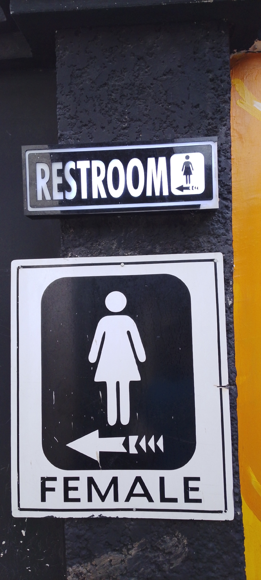 The image shows two signs mounted on a textured, dark gray concrete wall. The upper sign is a black rectangular sign with white lettering that reads "RESTROOM" followed by a white female silhouette inside a white square, with a white arrow pointing to the left. The lower sign is a larger black square sign with a white border. Inside the square, there is a large white female silhouette, and below it, a white arrow pointing to the left with triangular shapes on its shaft. Underneath the arrow, the word "FEMALE" is written in large white capital letters. To the right of the signs, a brightly painted yellow surface is visible, with some brush strokes suggesting it might be a mural or painted wall. The concrete wall appears to be an exterior surface. The scene does not provide any specific visual cues to pinpoint its location within the city of Howayi, Nigeria.
