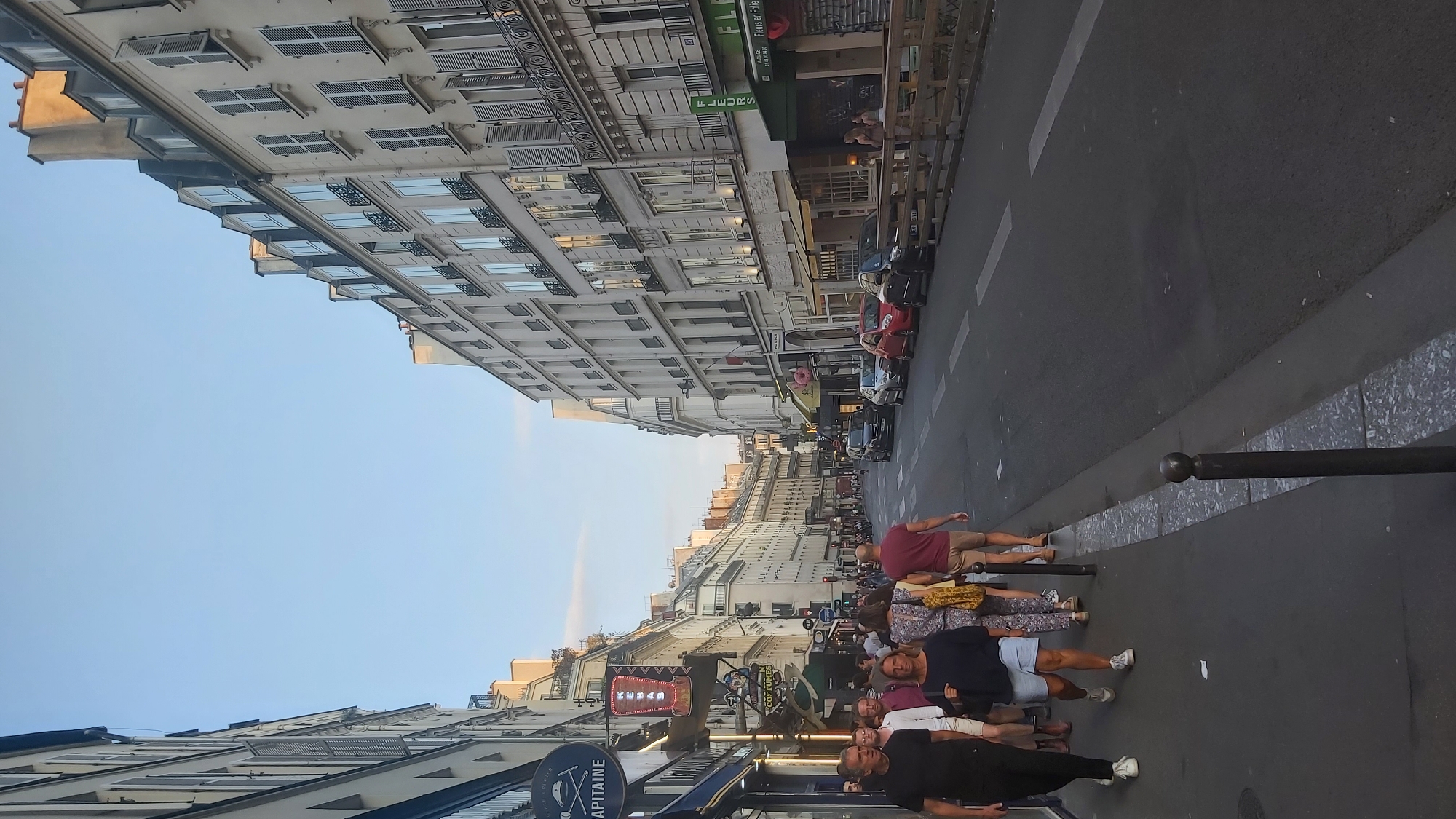 The image depicts a street scene in Paris, France, likely during the late afternoon or early evening, as indicated by the warm lighting and the long shadows. The perspective is from street level, looking down a relatively narrow street lined with multi-story buildings on both sides. These buildings appear to be typical Parisian architecture, characterized by their cream-colored facades, multiple windows with shutters, and pitched roofs with dormer windows.
On the left side of the street, several people are walking along the sidewalk. They are a diverse group, dressed in casual summer clothing. A man in a black t-shirt and dark pants is in the foreground, walking away from the camera. To his right, a woman in a dark blue top and light blue shorts is walking. Behind them, other pedestrians are visible, some carrying shopping bags. Several signs are mounted on the buildings on this side, including a circular sign with a skull and crossbones motif that says "CAPITAINE," and a neon sign that reads "KEBABS."
The street itself is paved and appears to be a one-way road with cars parked and driving on the right side. Several cars, including a black SUV, a red hatchback, and a dark-colored sedan, are visible. A wooden barrier runs along the right side of the street, separating the sidewalk from a row of shops. Some of these shops have awnings and storefront displays, including one with a sign for "FLEURS" (flowers). A "POLICE" sign is also visible on one of the buildings.
The sky is a clear, pale blue, with a few faint clouds visible towards the horizon. The overall impression is one of a bustling urban environment, likely a commercial street or a popular area for dining and shopping. The presence of pedestrians, shops, and cars suggests a lively atmosphere.