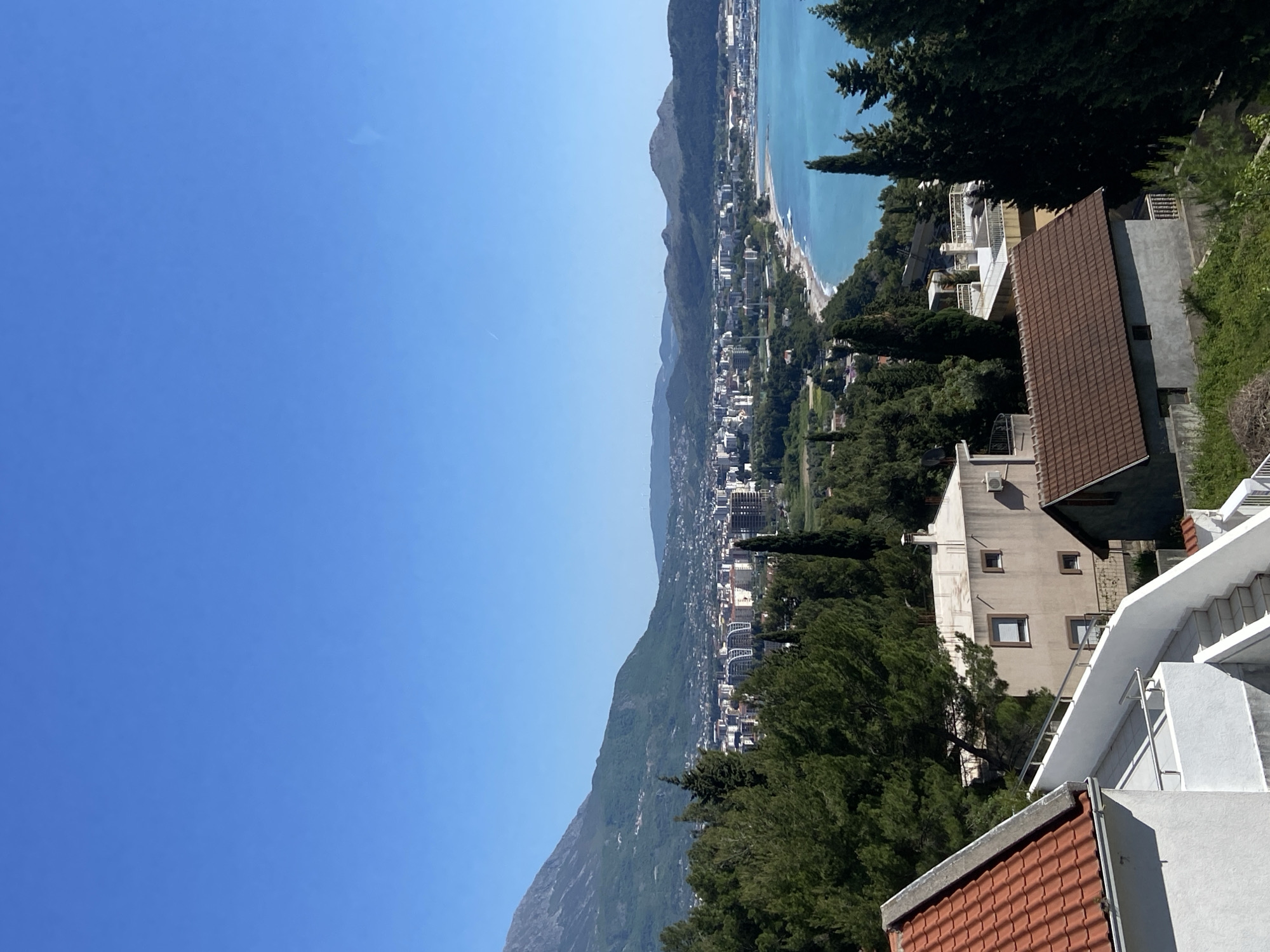 This is an elevated, panoramic view of the coastal town of Šušanj, Montenegro, under a clear, bright blue sky. The scene captures a wide expanse of the turquoise-blue Adriatic Sea to the right, a long coastline, and a dense urban area nestled between the sea and a prominent mountain range.
The sky is uniformly blue with only a very faint, wispy cloud or contrail visible in the upper left. The sea meets a narrow strip of beach that lines the coast. Along the coastline, numerous buildings form the town, characterized by a mix of multi-story structures and some with traditional reddish-brown tiled roofs, typical of Mediterranean architecture. The town extends inland, built into the lower slopes of the rugged mountains that rise steeply, forming a verdant backdrop.
In the immediate foreground, on the right side, are several residential buildings and abundant greenery. A house with a distinct red-tiled roof is prominent in the lower right. Behind it, a lighter-colored, multi-story building with multiple windows and balconies is visible. White outdoor stairs with railings ascend in the foreground, indicating a terraced hillside. Tall, dark green conifer trees, resembling cypresses, are interspersed throughout the landscape, adding to the lush appearance.
The image depicts a sunny day, likely mid-morning or early afternoon, with strong natural light. There are no people or specific activities visible, and no discernible text appears in the frame. The overall impression is one of a serene and picturesque Mediterranean coastal landscape.