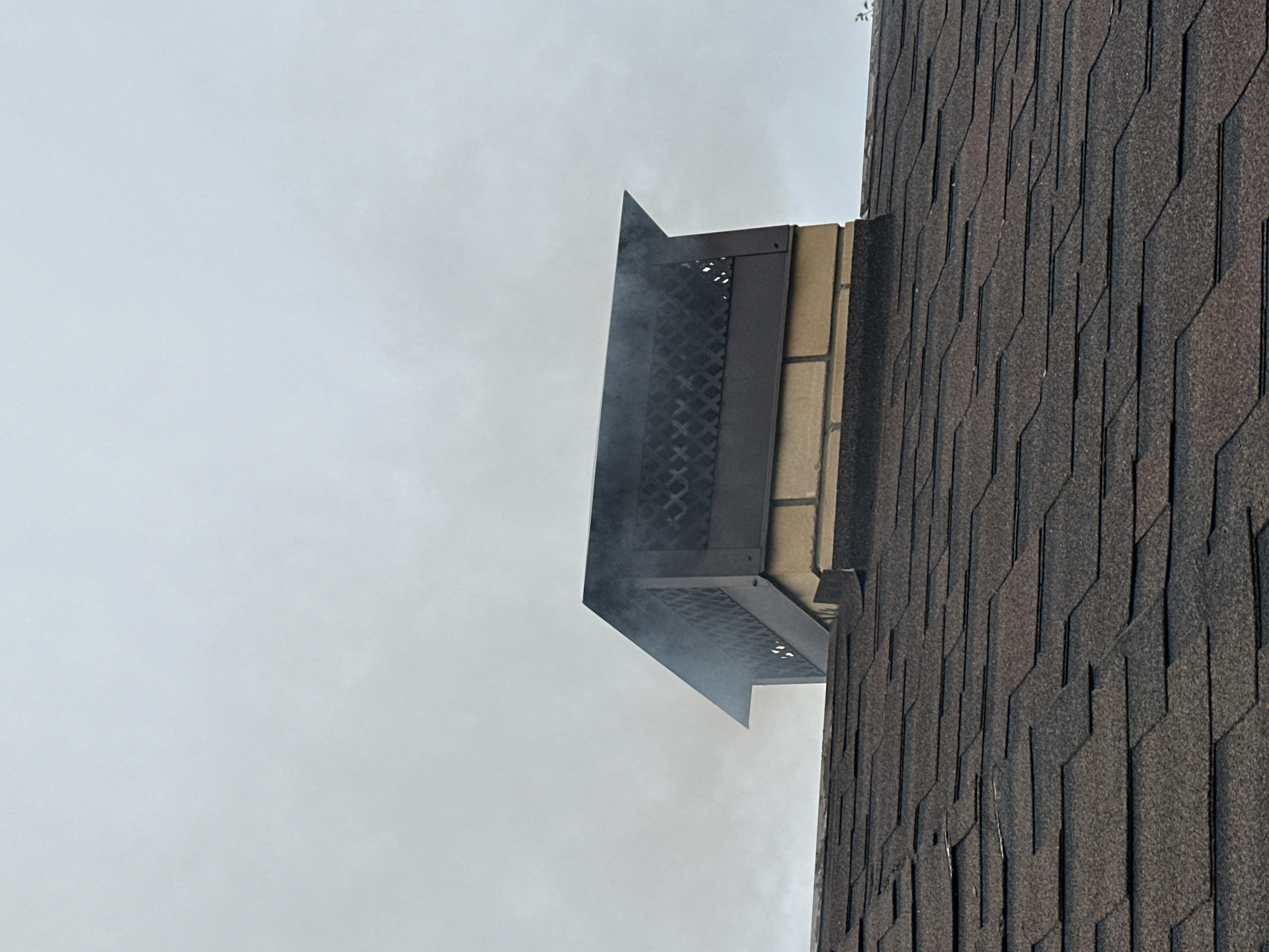 A close-up, low-angle shot captures the top of a residential building's roof, revealing a brick chimney with a dark metal cap. Light grey smoke is emanating from the top of the chimney, obscuring some of the decorative lattice on the cap. The roof is covered with dark brown asphalt shingles, some of which show signs of wear. The background is a uniformly grey, overcast sky. The scene appears to be in the city of Izobilnyy, Russia, on a day with poor visibility due to heavy cloud cover. There are no people or other significant objects visible in the frame.