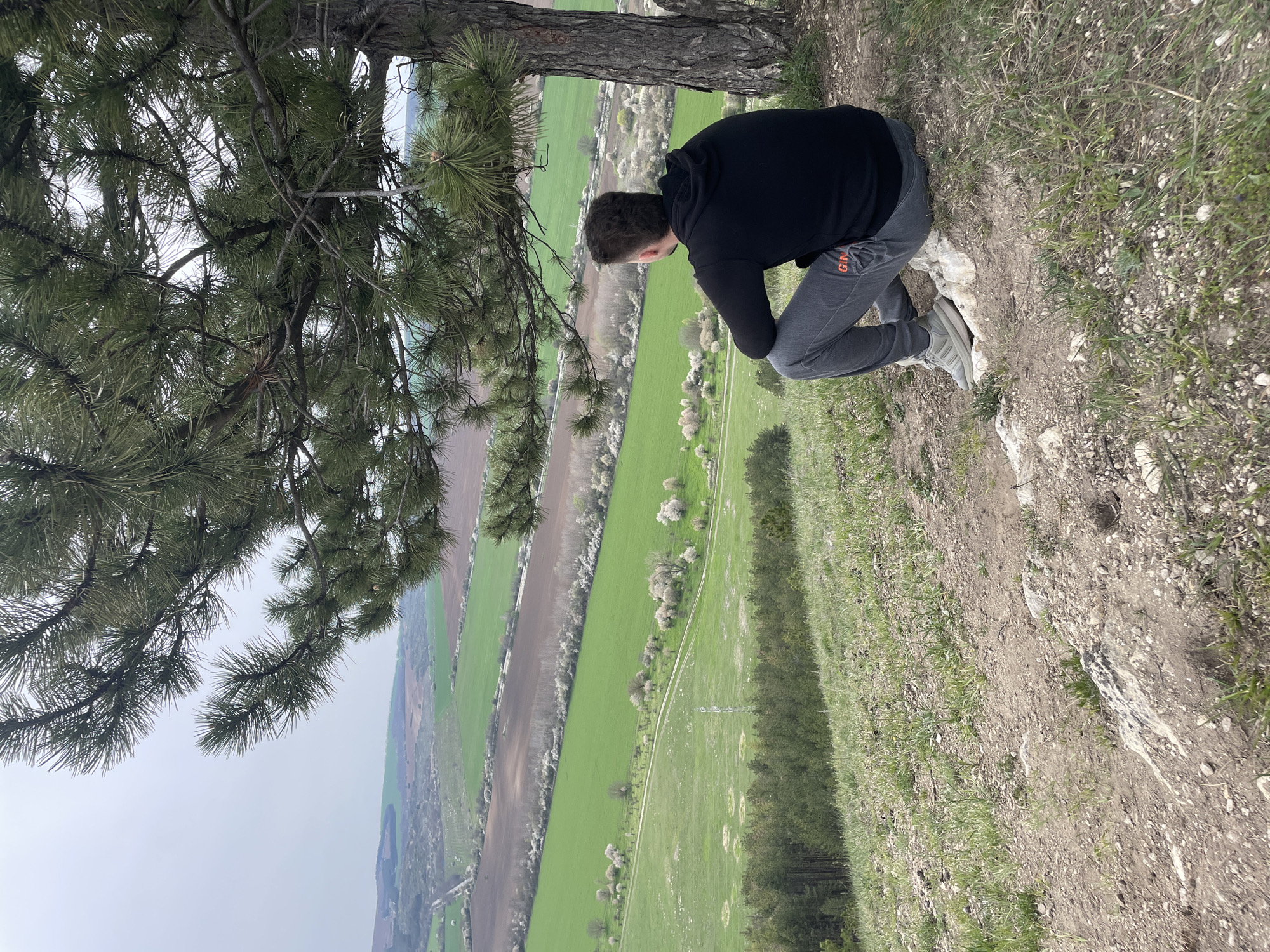 The image shows a person crouching near the edge of a cliff. The person is wearing a black hooded sweatshirt, grey sweatpants with orange lettering on the leg, and light-colored sneakers. They are looking down over the edge. A tree is situated on the cliff edge beside the person, with its branches extending out over the view. The background view is a landscape with fields of varying shades of green and brown. A dirt road runs through the fields. The sky is overcast and light grey. The cliff edge is covered in grass, dirt, and rocks. The location is Razgrad, Bulgaria.