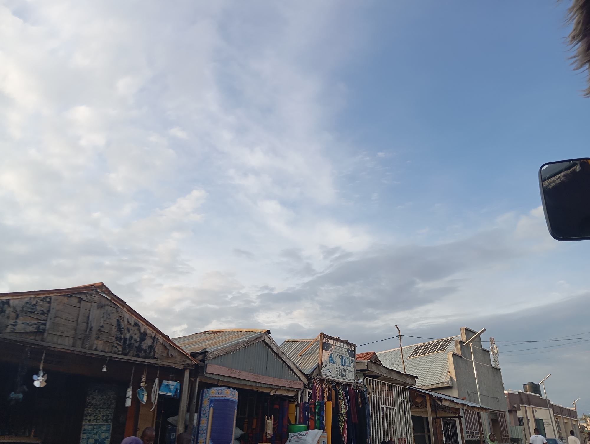 In Jalingo, Nigeria, a street scene unfolds with market stalls lining the right side of the image. The stalls feature corrugated metal roofs and wooden facades, displaying various goods such as rolled carpets, clothing, and other merchandise. One stall has a sign that reads "ABDULKARIM RAHUSA". The background reveals a mix of buildings, including a larger concrete structure with a sloped metal roof, and utility poles with wires crisscrossing the sky. The sky itself is a light blue with scattered white clouds, indicating daylight. A partial view of a vehicle's side mirror is visible in the upper right corner. Several people are present in the scene, walking along the street and in front of the market stalls.