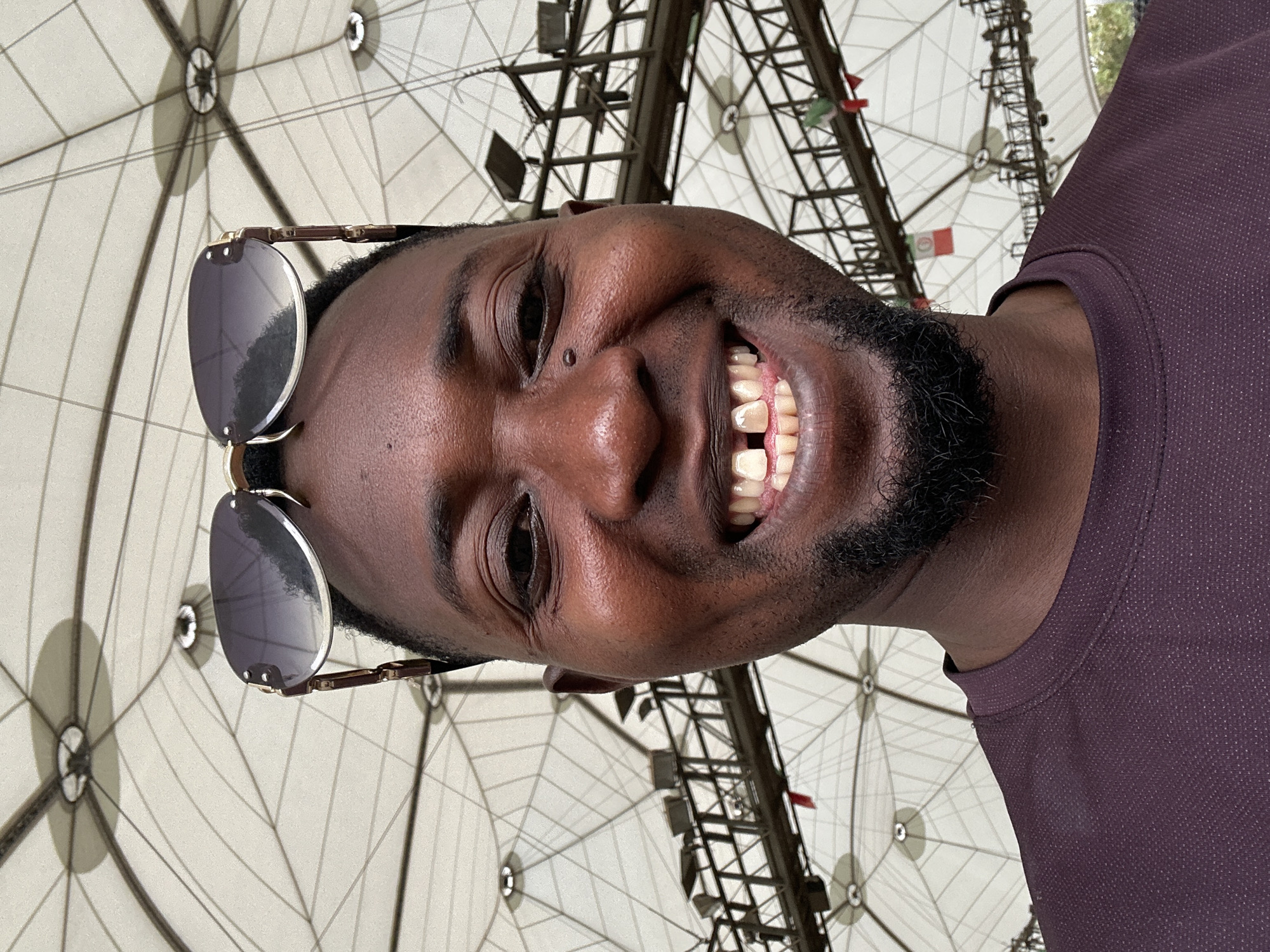The image shows a smiling man wearing sunglasses, with a beard, in front of a structure. The background structure appears to be made of metal and white fabric, and has a complex, geometric design. Small flags are hanging from the structure. The man is wearing a dark-colored, possibly purple, short-sleeved shirt. The location is unknown.