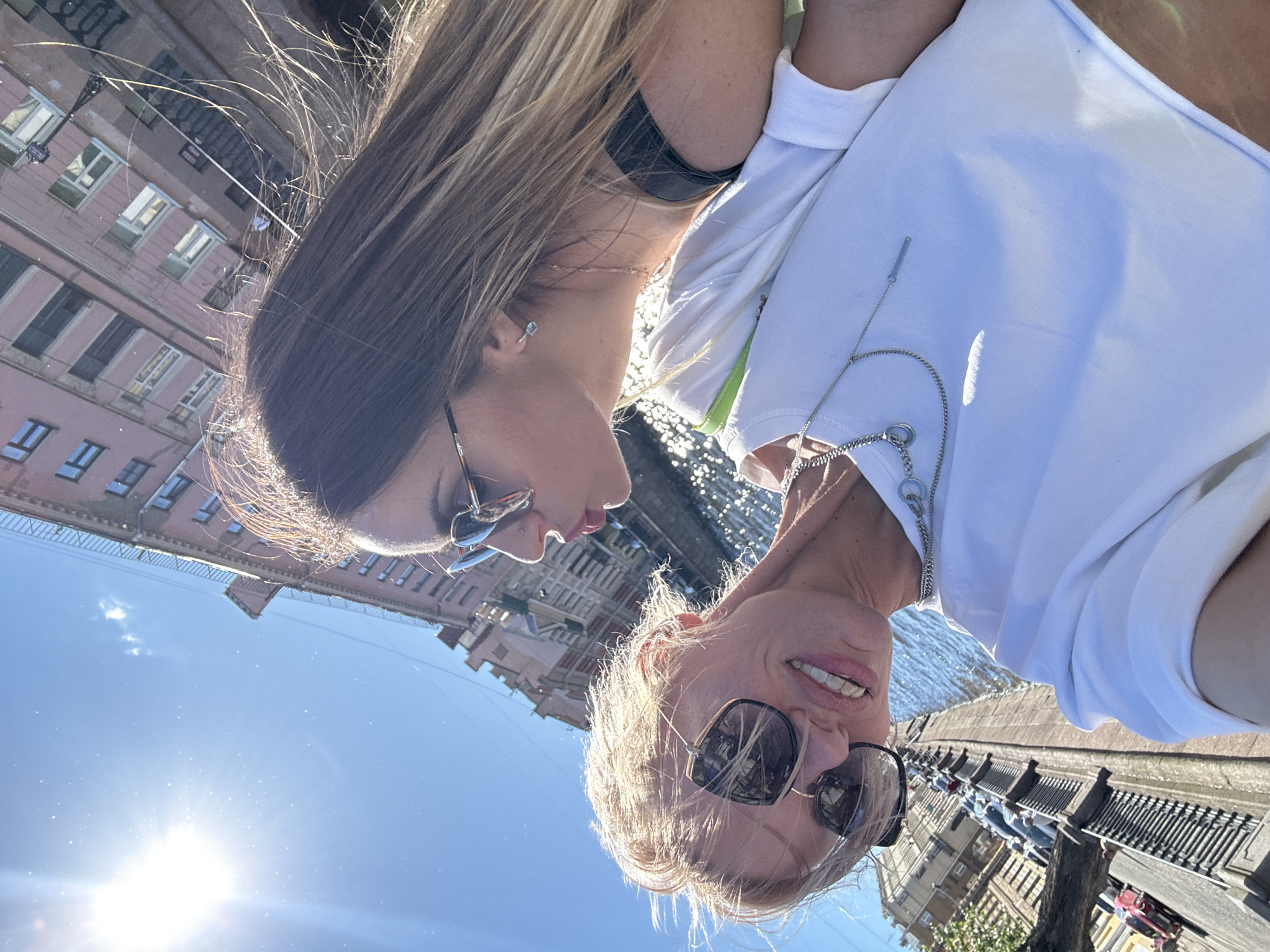 In Saint Petersburg, Russia, on a bright, sunny day, two women are taking a selfie by a canal. The woman in the foreground, on the left, has short, blonde hair and is wearing large, dark sunglasses and a white t-shirt. She is smiling and looking at the camera. A silver necklace with a bar pendant is visible. The woman on the right has long, brown hair and is also wearing sunglasses. She is looking up at the other woman, and they appear to be close. Her hair is illuminated by the sun. In the background, there is a canal with sparkling water, and across the canal, there are several multi-story buildings with pink facades and arched windows. The architecture suggests a historic European city. The sun is high in the sky, casting bright light and some lens flare onto the image. The overall atmosphere is one of leisure and enjoyment.