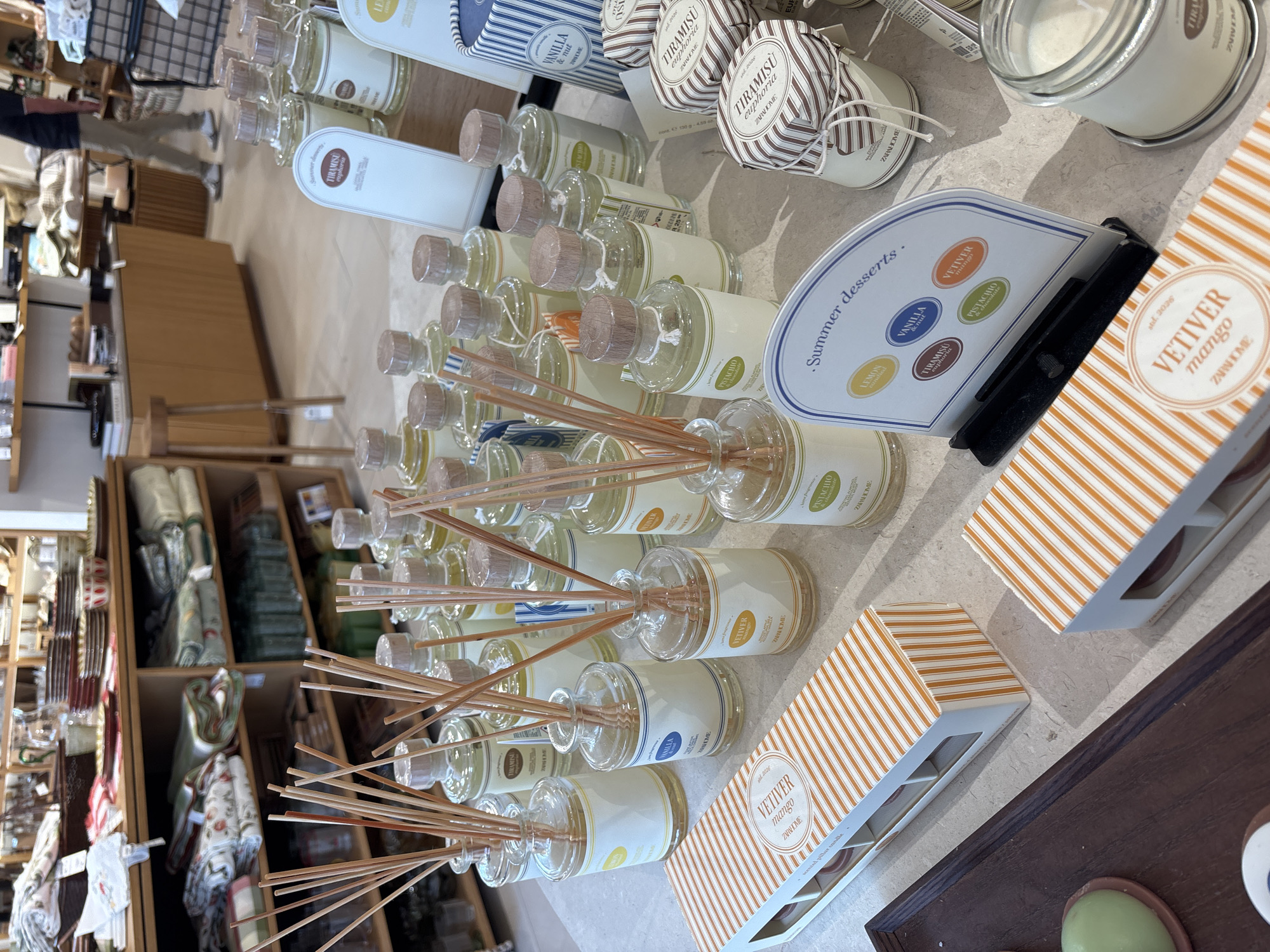 The image shows a display of home fragrance products, likely in a retail store. In the foreground, there are several glass bottles with wooden stoppers and reeds, indicating reed diffusers. These bottles have labels with various scent names, such as "VETIVER MANGO," "VANILLA & NUT," and "TIRAMISU EUPHORIA." A box with an orange and white striped pattern is also visible, labeled "VETIVER MANGO scented pillar candle." To the right, there are jars with striped lids, also labeled with scent names. A sign with the heading "Summer desserts" displays several oval labels corresponding to different scents. In the background, shelves are stocked with various home goods, including linens and tableware. The lighting suggests an indoor retail environment. The city of Orihuela, Spain, is not depicted or implied by any elements in the image. There are no people clearly visible in a way that allows for detailed description. The overall scene is one of organized product display within a store.
