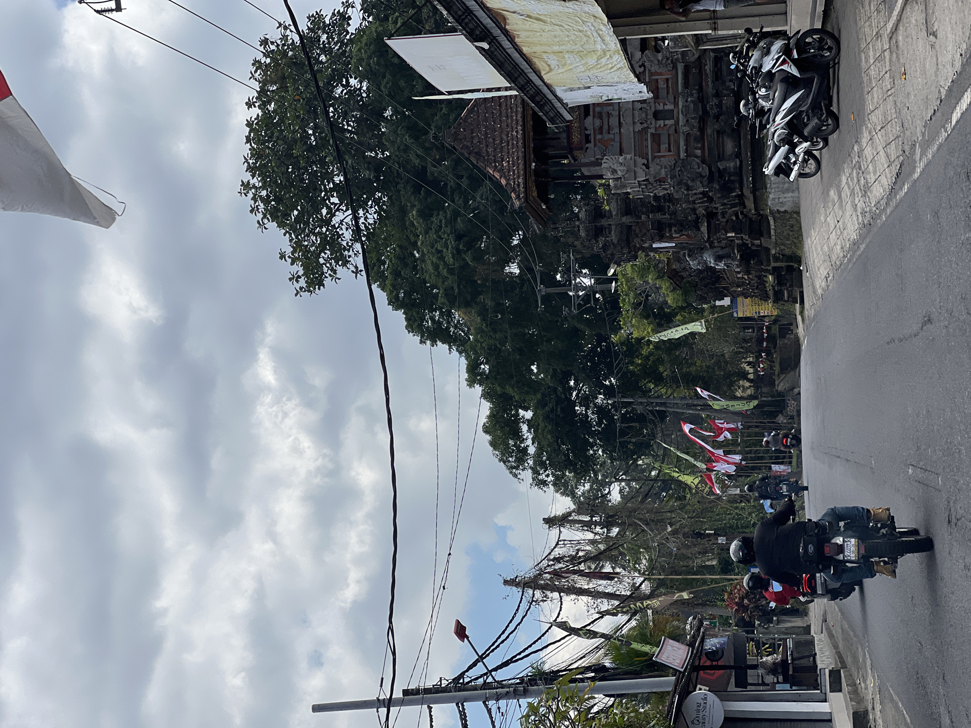 The image depicts a street scene in Payangan, Indonesia, under a cloudy sky. The foreground shows a paved road with several motorcycles. A person wearing a black jacket and helmet is riding a motorcycle in the center, with another person in a red shirt on a motorcycle ahead of them. The left side of the road features a building with signage, including "Oming Beauty Studio". Further down the road, there are more motorcycles and flags, including red and white ones. The right side of the road has a traditional Balinese temple structure with intricate stone carvings and a tiled roof, with several parked motorcycles in front of it. A large tree with dense foliage is visible behind the temple. Overhead, numerous electrical wires are strung across the street, supported by utility poles. A large white and red flag is flying in the upper right corner of the image. The overall impression is of a moderately busy street in a tropical environment, likely a tourist area given the temple and the presence of flags.
