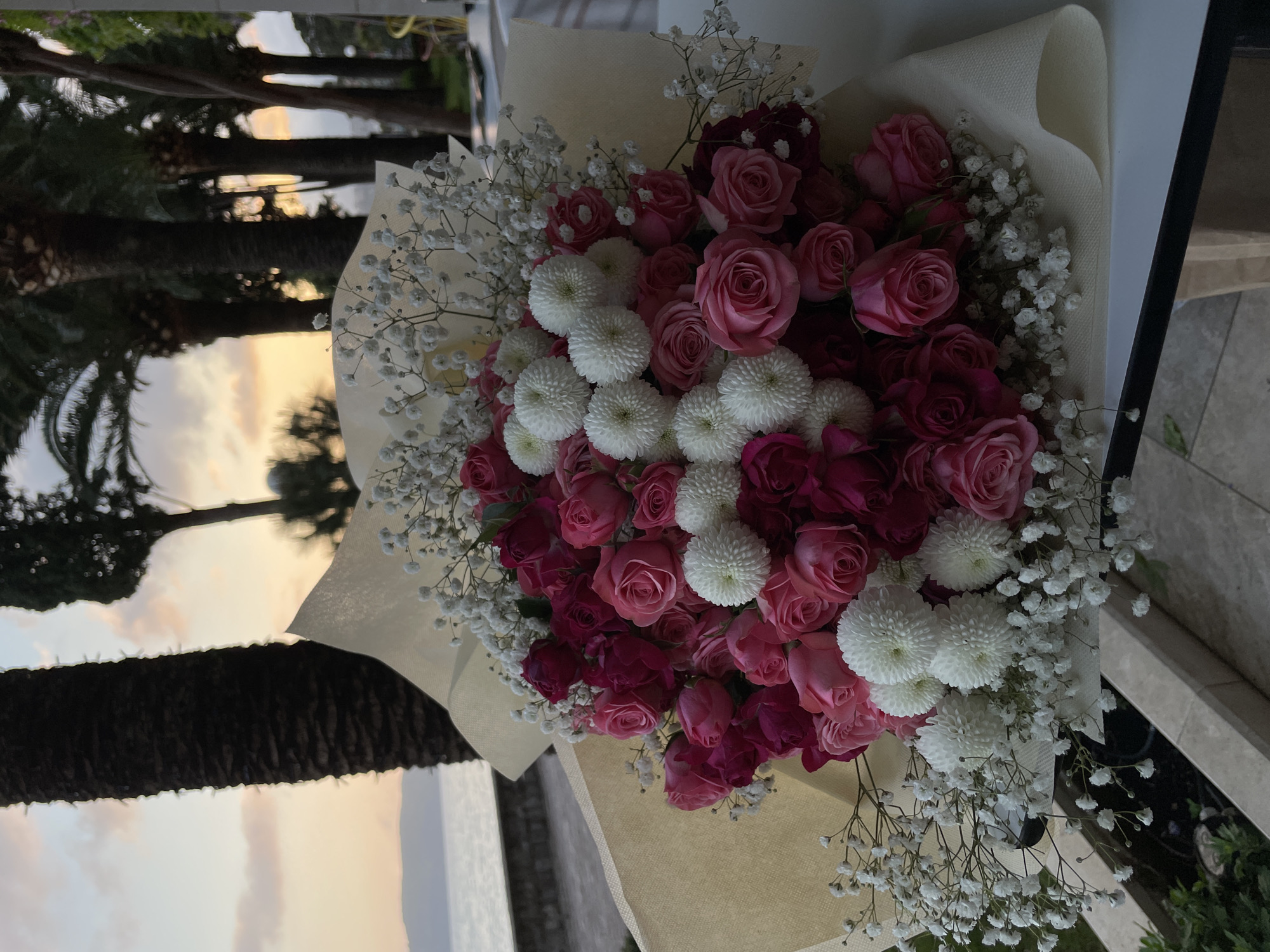 The image displays a large, vibrant bouquet of flowers set against an outdoor background, captured in soft, low light conditions. The bouquet primarily features numerous roses in shades ranging from delicate pink to deep magenta. These are artfully mixed with clusters of small, spherical white flowers, resembling pompom chrysanthemums, and delicate sprigs of white baby's breath (Gypsophila) that fill the gaps and add texture. The arrangement is wrapped in a light-colored, possibly cream or beige, textured paper or fabric.
The background reveals a serene outdoor scene at what appears to be either sunrise or sunset. Tall palm trees are silhouetted against a sky that transitions from warm, soft orange and yellow hues near the horizon to a muted blue-grey higher up. Subtle reflections are visible, suggesting the presence of glass or a calm body of water, hinting at a coastal or waterfront location. The presence of palm trees and a scenic view is consistent with a Mediterranean setting like Rafailovići, Montenegro. No people or visible text are present, focusing the composition on the beauty of the floral arrangement and its tranquil natural surroundings.