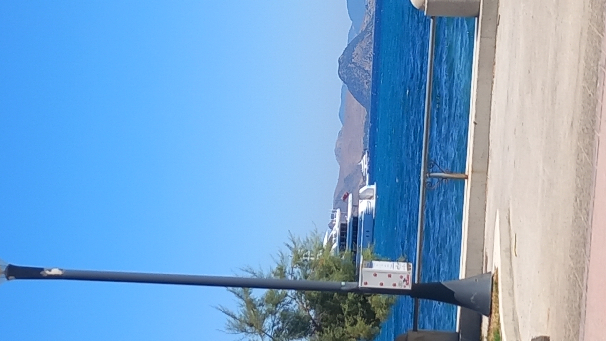 The image shows a bright, sunny day with a clear blue sky over the Aegean Sea. In the foreground, a dark grey lamppost stands on the left, with a notice attached to it. A low concrete wall with a metal railing separates a paved area from the sea. A lush green tree with sparse foliage is visible behind the lamppost. In the middle ground, several white yachts are anchored in the vibrant blue water, the largest of which is prominent and features multiple decks and a red flag. In the background, a chain of islands with rugged, brown hills and a dark, conical peak are visible against the horizon. The overall scene depicts a picturesque coastal view in the city of Kos, Greece, likely a harbor or waterfront area.