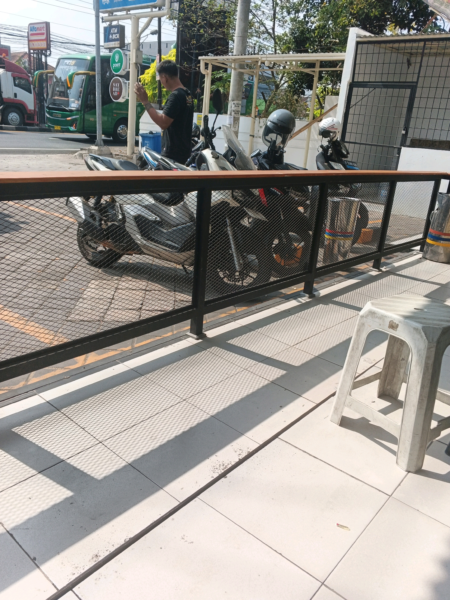 In Genuk Barat, Indonesia, a man is seen standing near a bus and several motorcycles. He is looking at his phone, holding it in his right hand. He is wearing a dark t-shirt and appears to be of average build. The man is positioned behind a black metal fence. In the background, a green bus with "PUNCAK" written on it is parked, along with a white truck. Various signs are visible, including one for "Alfamart" and "ATM BCA." Several motorcycles, two of which have helmets on them, are parked behind the fence. The area appears to be a street or a parking lot, with a tiled ground surface. Sunlight casts shadows from the fence and the motorcycles onto the ground. A white plastic stool is present in the foreground, to the right of the frame.