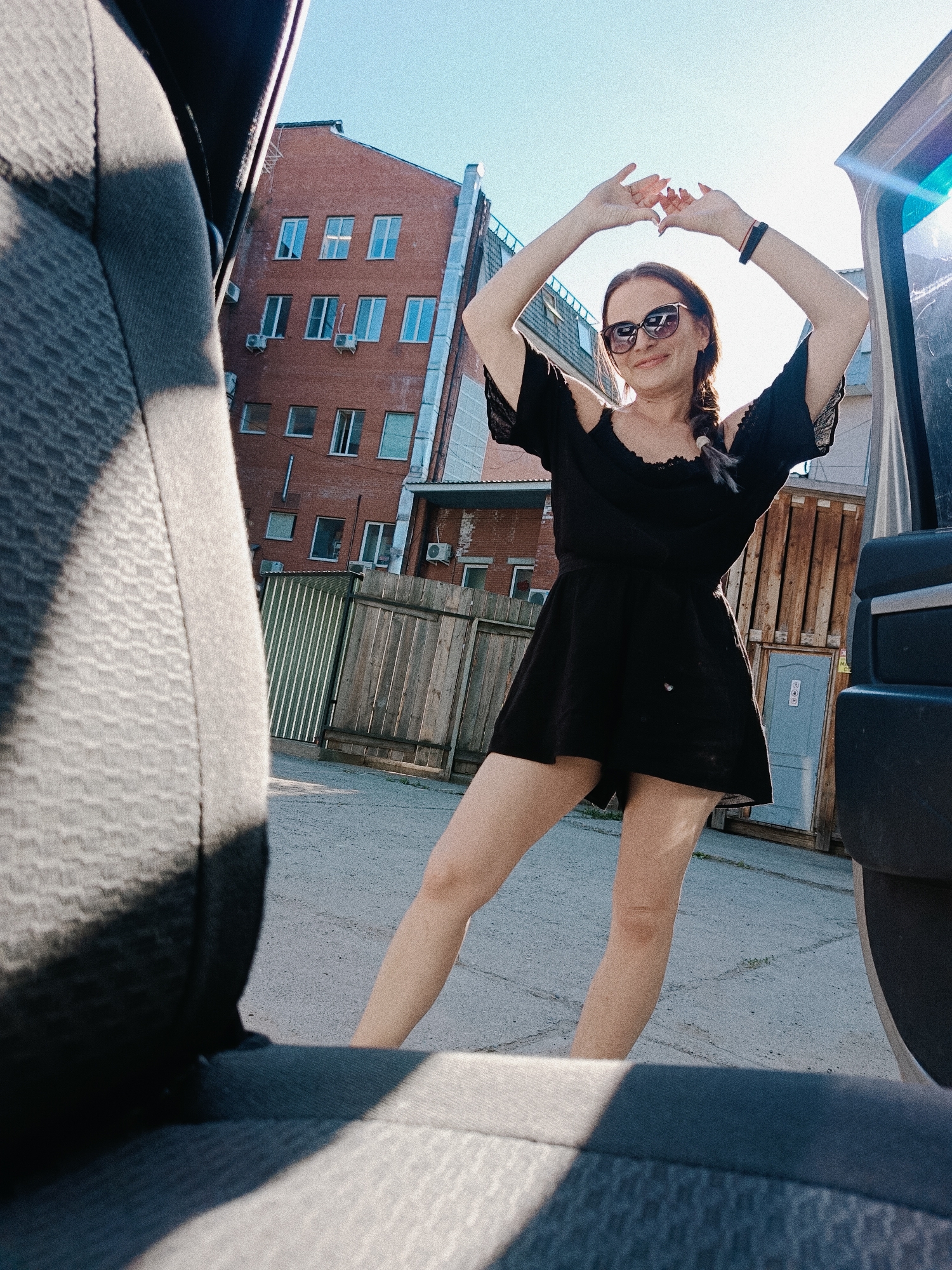 A fair-skinned woman in a black dress and sunglasses stands with her arms raised near a dark-colored van. The woman has her hair braided and is smiling. The background includes a brick building, a wooden fence, a portable toilet, and an asphalt surface. The photo is taken from inside a vehicle, as a gray car seat and part of the vehicle frame are visible in the foreground. The location is Chaikovsky, Russia.