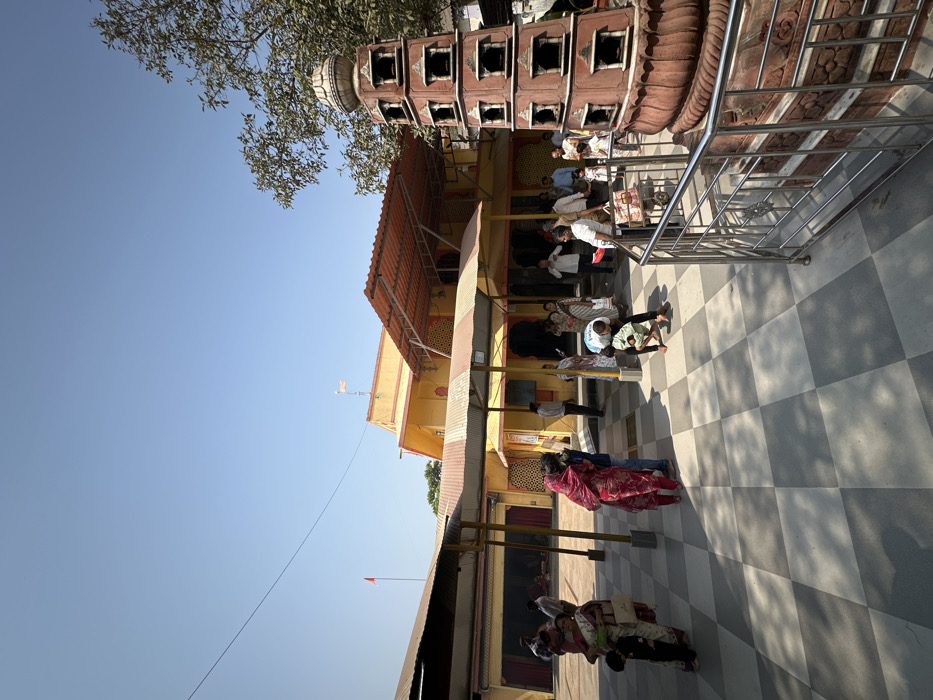 A vibrant temple scene unfolds with visitors dressed in casual attire, including a woman in a striking red saree. The temple, bathed in sunlight, exhibits a yellow facade with an orange rooftop. Ornate brickwork adorns a side structure, possibly a tower. A checkered tile floor provides a clear view of the space where people are standing or sitting. A clear blue sky is visible overhead.