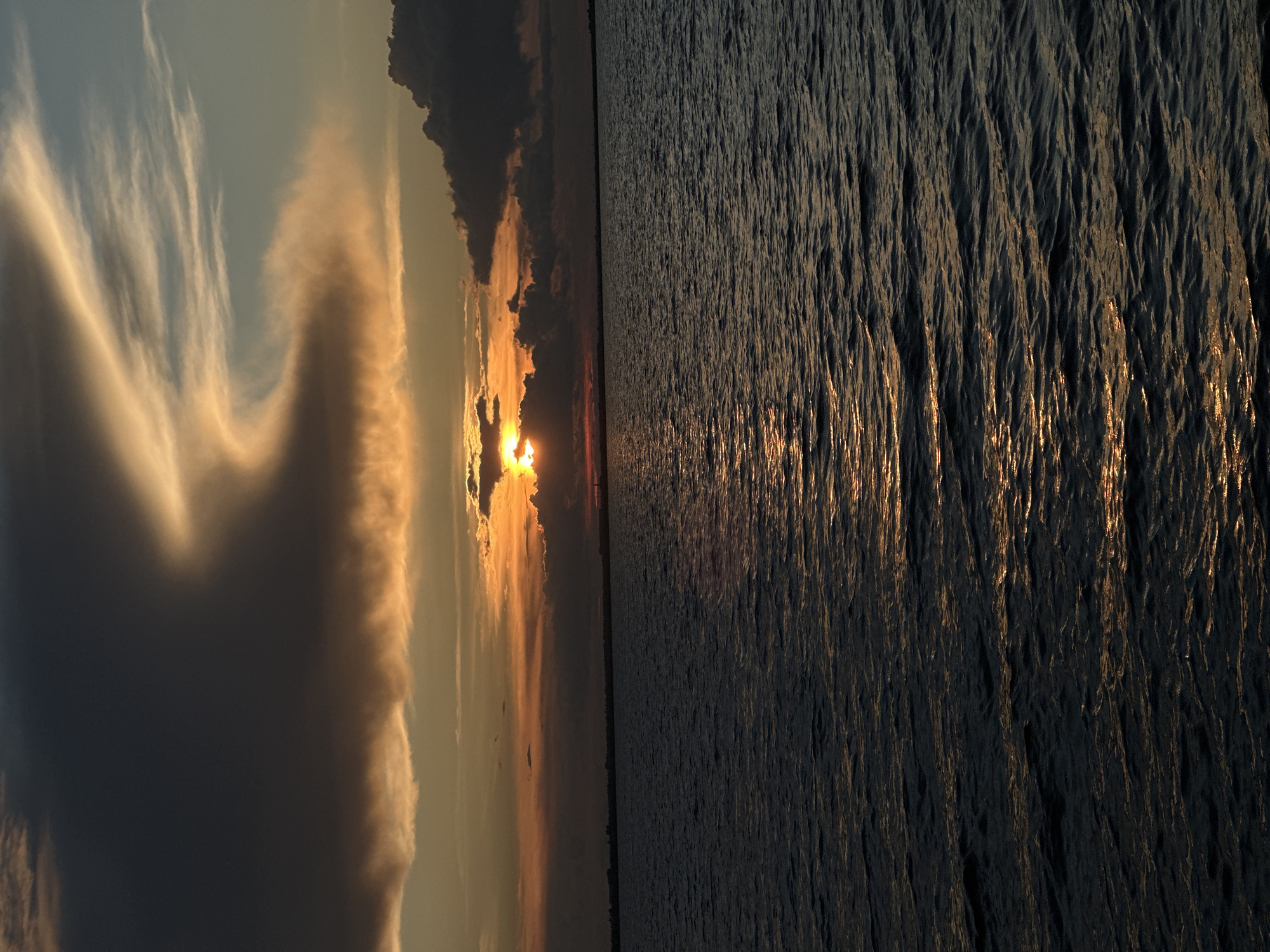 The image depicts a sunset over a body of water. The sky is filled with dramatic clouds, ranging from dark, ominous masses to wispy, illuminated streaks. The sun is partially obscured by clouds, casting a warm, orange glow across the horizon. The water in the foreground is dark and choppy, reflecting the colors of the sunset in shimmering patterns. A faint, dark line of land is visible in the distance, suggesting the presence of a shoreline or city. The overall impression is one of a serene yet powerful natural display. The location in the city of Kokshetau, Kazakhstan, is not discernible from the image itself, as there are no identifying landmarks or specific features that tie it to that particular place.