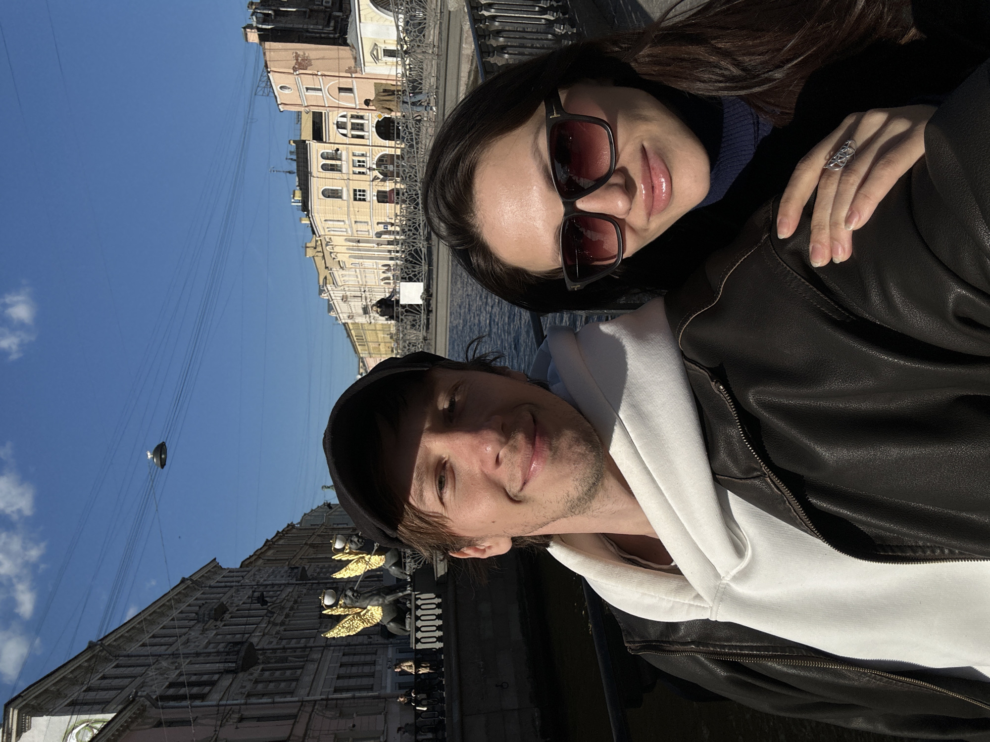 A man and a woman are posing for a selfie-style photograph in Saint Petersburg, Russia. The woman has long dark hair and is wearing sunglasses. She is also wearing a dark-colored turtleneck sweater. A silver ring is visible on her right hand. The man is wearing a baseball cap, a white hooded sweatshirt, and a dark leather jacket. Behind them, there is a canal, a bridge, and buildings typical of Saint Petersburg architecture. Golden statues or architectural details are visible on one of the buildings. The sky is blue and clear.