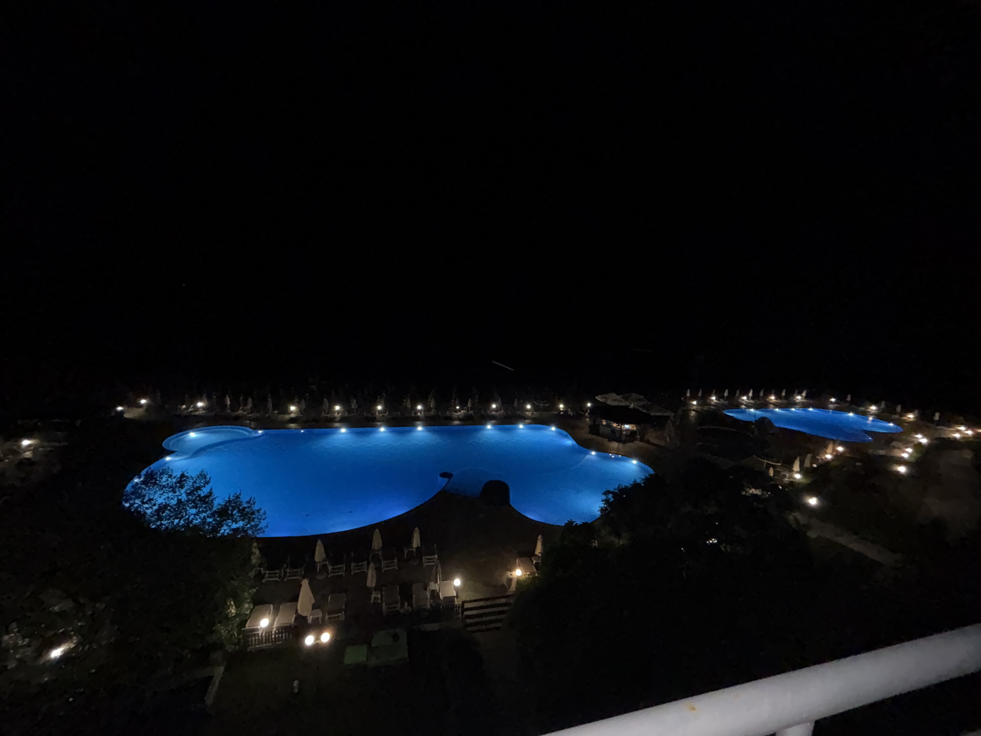 This is a high-angle, nighttime photograph of a resort with multiple illuminated swimming pools. The main pool is large and irregularly shaped, with bright blue lighting. A smaller, rectangular pool is visible to the right, also illuminated. The surrounding area is dark but dotted with lights, suggesting pathways and building lights. Rows of lounge chairs and umbrellas are visible, some of which are illuminated by nearby lights. Trees and vegetation are present, appearing dark against the lit pools and sky. The image captures a tranquil, nocturnal resort scene, likely in Varna, Bulgaria, given the context.