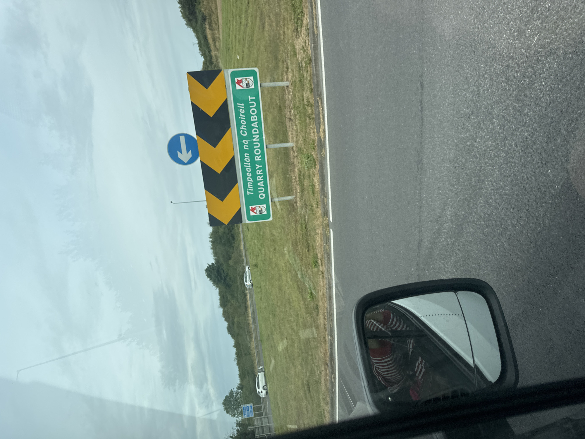 The image, taken from a vehicle, shows a road sign with the text "Timpeallán na Choiréil QUARRY ROUNDABOUT" in English and Irish. The sign is green with white lettering and has two crests on either side. Above the sign is a larger chevron sign with yellow and black markings, indicating a traffic diversion. A blue circular sign with a white left-pointing arrow is positioned above the chevron sign. In the background, there is a grassy embankment with some trees and two white cars visible on the road. The sky is overcast. A reflection of a person wearing a striped shirt is visible in the side-view mirror of the vehicle. The scene appears to be in Ireland, likely on a highway approaching a roundabout.