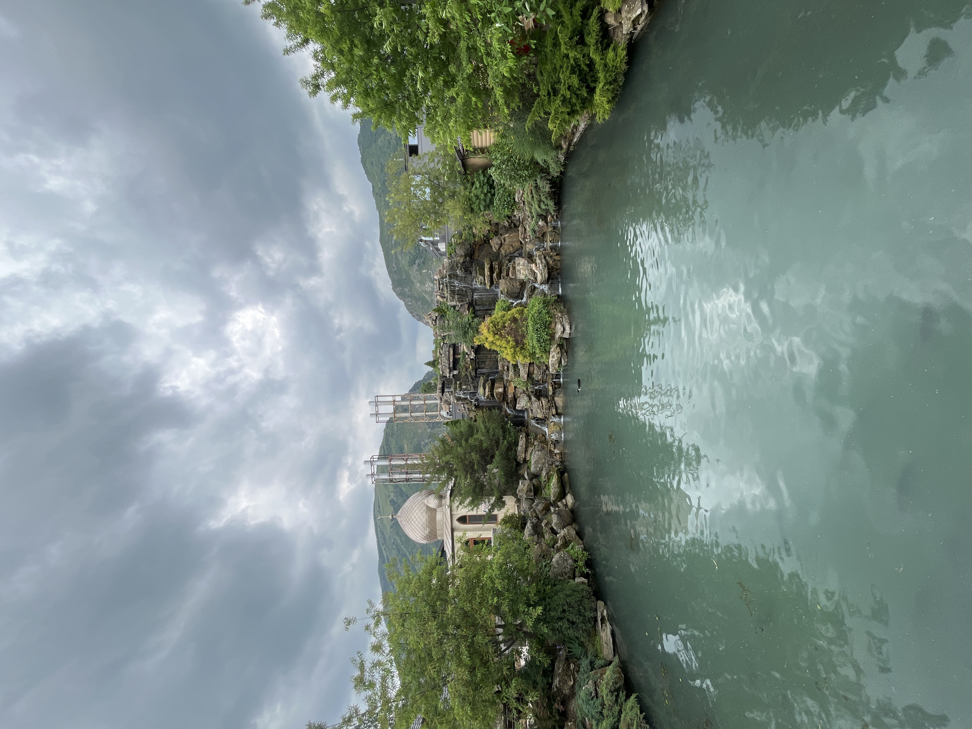 The image depicts a pond or narrow lake, its surface reflecting the overcast sky. Along one side of the water, a rocky bank is overgrown with vegetation, including trees and smaller plants. A building with a dome is visible among the trees on the rocky bank. Behind the building, a mountain rises, its peak obscured by clouds. A structure resembling scaffolding is also visible behind the building. The scene appears to be set in a natural or landscaped environment, despite the presence of the scaffolding. The location is in the city of Miatli, Russia.