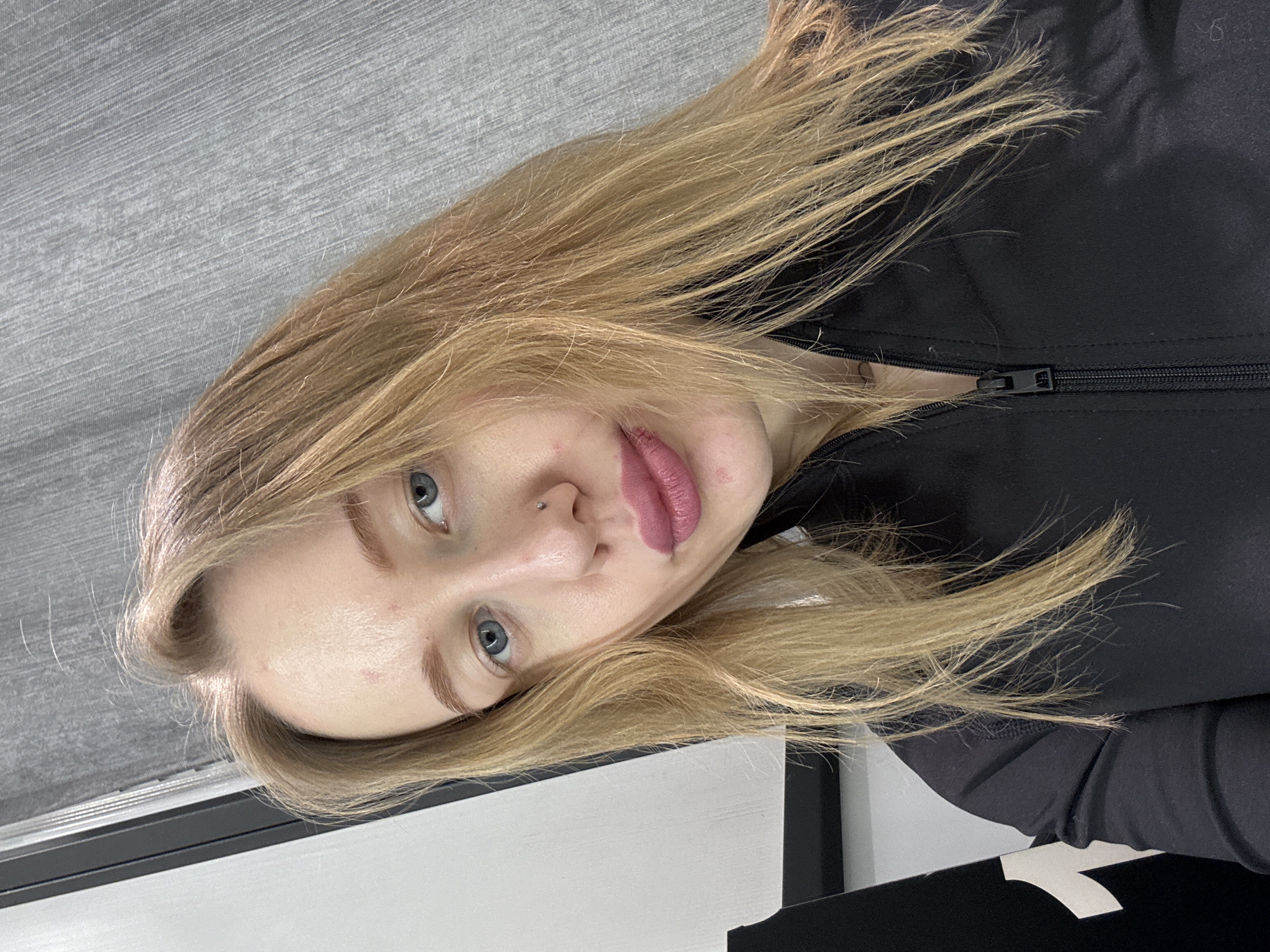A fair-skinned person with blonde hair is seen wearing a black zippered garment. The person has blue eyes, light eyebrows, and a nose piercing. The background consists of a gray surface and a white and black structure. There is no information to indicate this photo was taken in Rostov-on-Don, Russia.