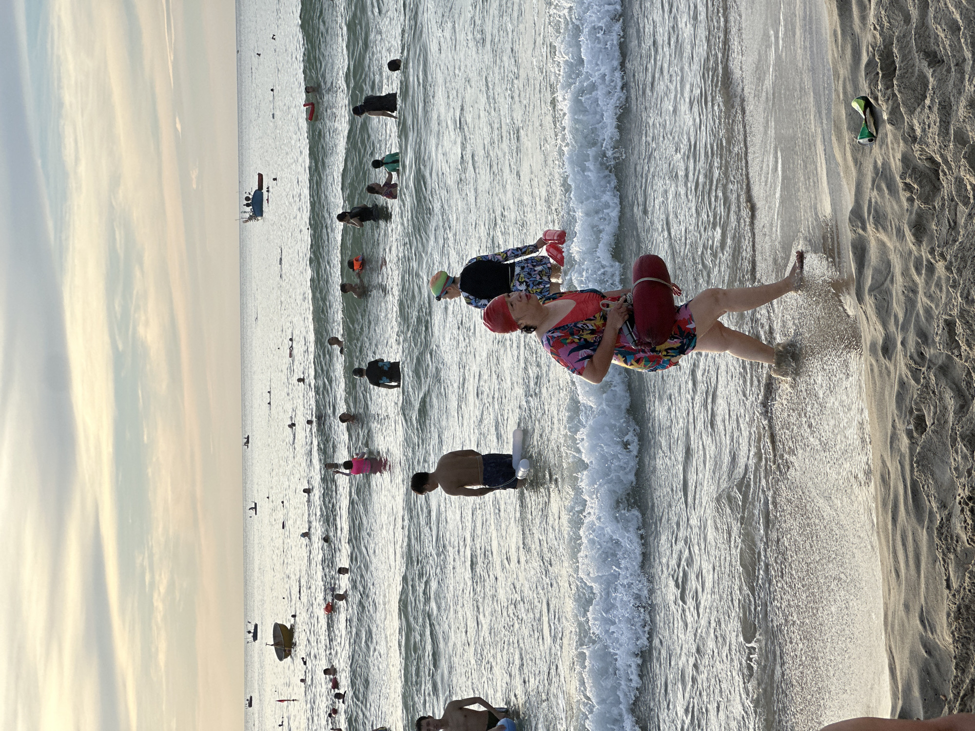 The image depicts a crowded beach scene in Đà Nẵng, Vietnam, likely during sunset given the warm, diffused light and the hazy sky. The ocean is populated with numerous swimmers and several small boats are visible on the horizon. In the foreground, a woman in a colorful swimsuit and red headscarf is walking towards the viewer, carrying a red life buoy. Another woman, wearing a black and patterned swimsuit and a swim cap, is also present in the water. Several other individuals are swimming and wading in the ocean, with some appearing to be playing or relaxing. The beach itself has soft sand with a few small dunes. A discarded aluminum can lies on the sand in the lower right portion of the frame, suggesting recent human activity. The overall impression is of a lively and active beach environment.