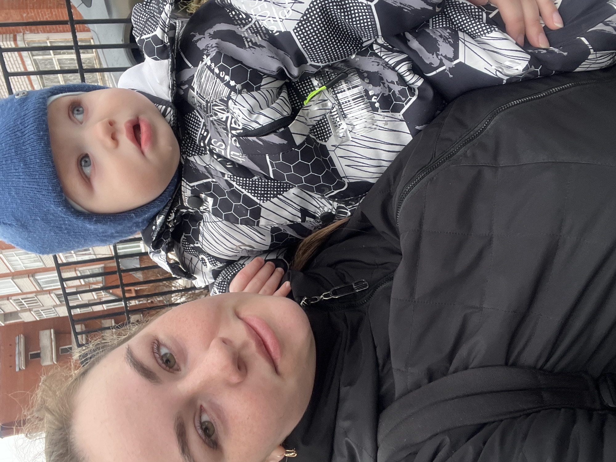 A woman and a young child are outside in what appears to be an urban environment. The woman is wearing a dark-colored quilted jacket, and the child, wearing a blue knit cap, is dressed in a patterned, possibly winter, jacket. The building in the background has multiple windows and appears to be residential or commercial. A metal railing is visible in the foreground. The image was taken in the city of Chaikovsky, Russia.