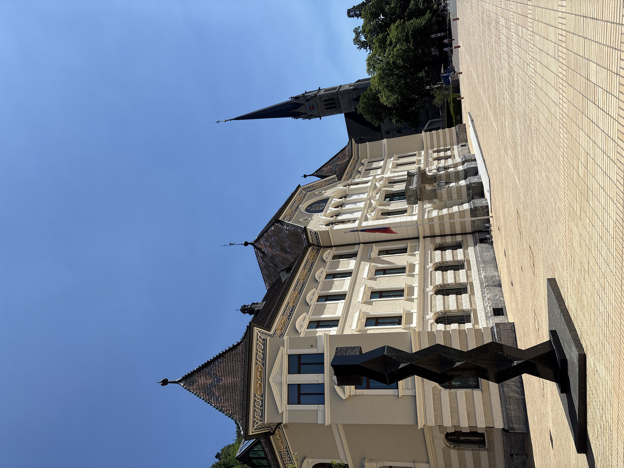 The image shows a sunny day in Vaduz, Liechtenstein. In the foreground, a black abstract sculpture stands on a stone base in a paved plaza. To the left, a light-colored building with ornate detailing and a steep, tiled roof is visible. A flagpole with the flag of Liechtenstein is mounted on the building. Behind this building, a tall church spire with a clock face rises into the clear blue sky. Lush green trees are scattered throughout the background, and a few people can be seen walking in the distance. The plaza is paved with light-colored square tiles arranged in a patterned grid.