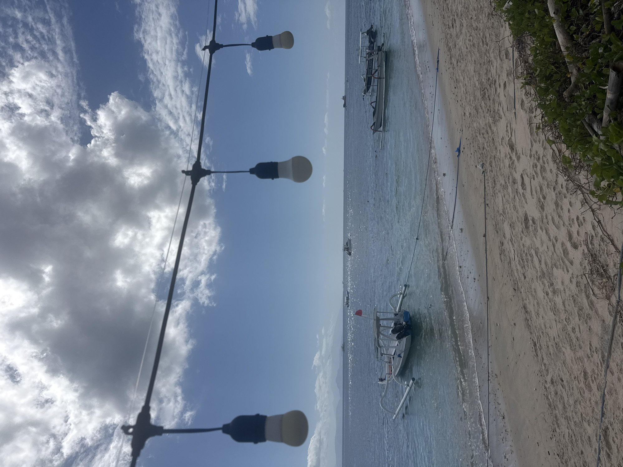 The image depicts a serene beach scene in Nusa Penida, Indonesia. The foreground shows a sandy beach with some green foliage and driftwood on the right side. A series of lights hang on a wire across the top of the frame. The middle ground features the calm, blue ocean with several boats anchored. The largest boat, a white outrigger canoe, is prominent in the center-left of the image, equipped with an outboard motor and a small red flag. Other boats, including two smaller outrigger canoes, are visible further out in the water. The background reveals a hazy horizon with distant landmasses and a bright blue sky dotted with fluffy white and gray clouds. The water appears clear and shallow near the shore, with gentle waves lapping the beach.