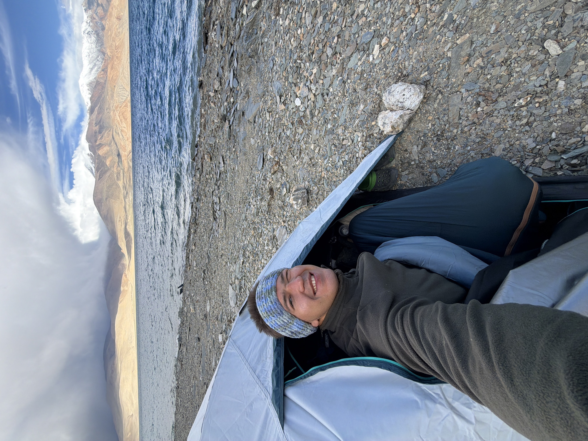 A man, wearing a dark green fleece jacket and a blue and white knitted headband, is pictured from a first-person perspective, lying inside a light blue tent. He is smiling and looking at the camera. The tent is pitched on a rocky shore beside a body of water. In the background, there is a vast expanse of water, a barren, tan-colored mountainous landscape under a cloudy sky, and further in the distance, snow-capped mountains. The overall scene suggests a camping trip in a remote, mountainous region.