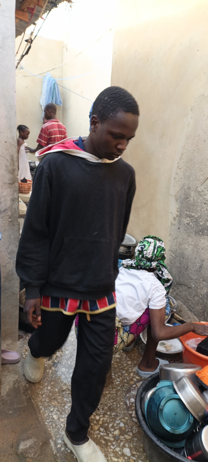 The image captures a candid moment in a narrow outdoor corridor or alleyway, likely within a residential compound in Kaura Goje, Nigeria. The walls are light-colored and appear to be made of concrete or mud brick, with a corrugated metal roof visible in the upper left corner.
In the foreground, a young Black man walks towards the left side of the frame. He is dressed in a black hooded sweatshirt with a red and white lining visible in the hood, worn over a blue collared shirt. A distinctive band of red, blue, and yellow striped fabric is visible beneath the hem of his sweatshirt, possibly an underlying garment. He wears black pants and light-colored, slip-on shoes or clogs. His head is slightly bowed, and his expression is neutral.
To the right, partially obscured by the foreground man, a woman is seen from behind, squatting or kneeling. She is engaged in washing dishes, immersing her hands in an orange basin. She wears a white t-shirt, a vibrantly patterned skirt or wrapper in green, pink, and yellow, and a matching green and white patterned head covering. Various metal and plastic bowls are visible in a larger black basin next to her.
Further in the background, to the left, another man in a red and white striped shirt stands near a young child. The child is wrapped in a white cloth or towel and stands beside a brown woven basket. A light blue piece of cloth hangs on a line strung across the alleyway. The ground is wet and appears to be a mix of concrete and pebbles. The bright lighting suggests it is daytime. There is no visible text in the image. The scene depicts routine domestic activities in a bustling outdoor setting.